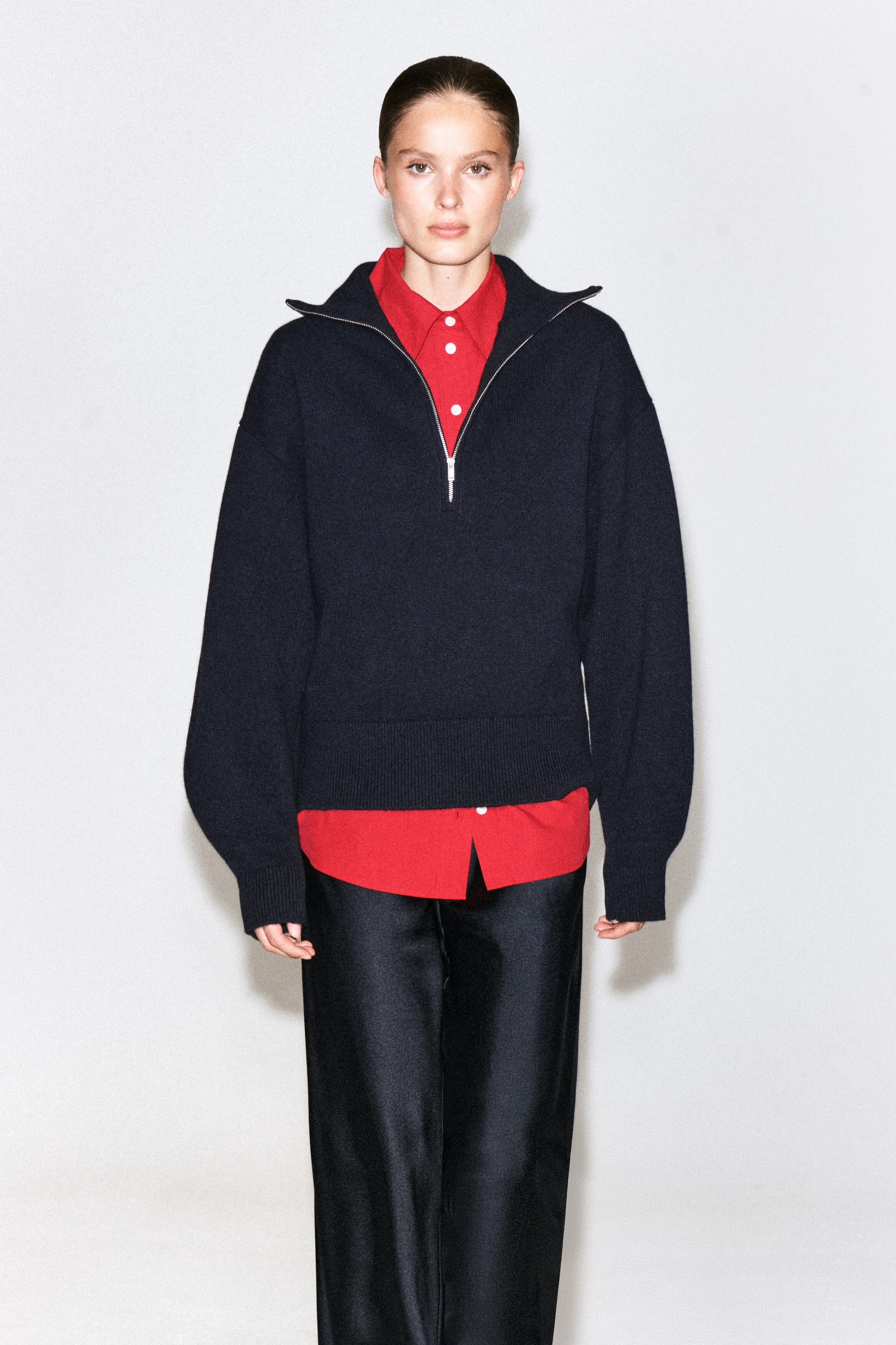 A dark navy knit half-zip jumper is being worn by the model, featuring a silver zip, relaxed sleeves, and a ribbed hem and cuffs. The jumper is layered over a bright red collared shirt and paired with black trousers.