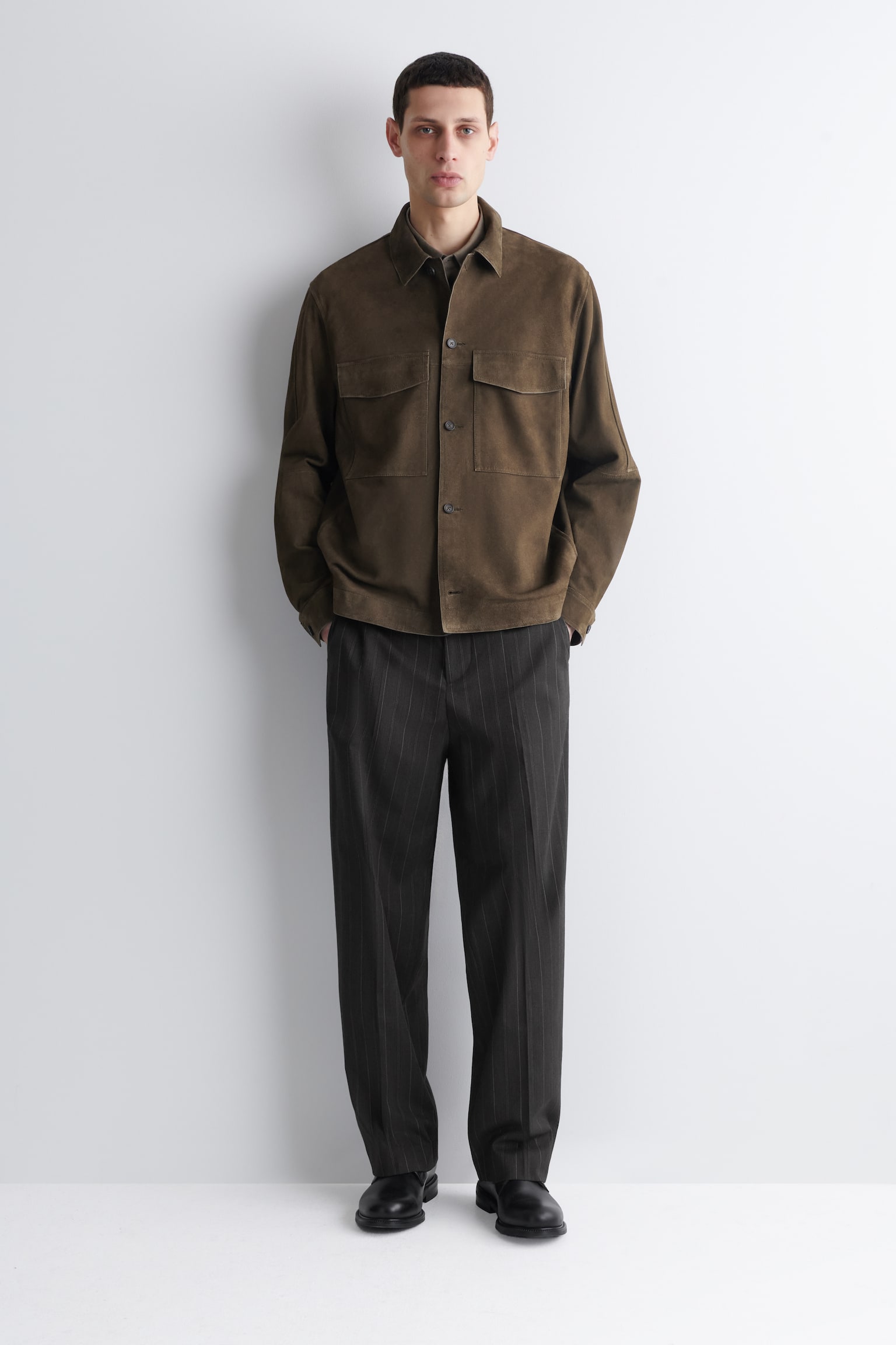 Une veste-chemise en daim vert olive foncé, avec un col pointu et deux poches à rabat sur la poitrine, est portée sur une chemise à col sombre. Elle est associée à un pantalon à rayures fines gris foncé, avec une coupe droite et décontractée, ainsi que des chaussures noires à bout uni, tous orientés vers l'avant. - 1