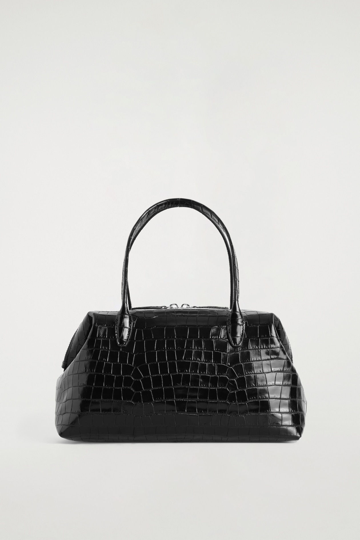 A black croc-embossed leather bowling bag is shown from the front, highlighting its structured, sculptural shape with softly pinched sides. It features two curved top handles and a silver-toned two-way zip fastening along the top.