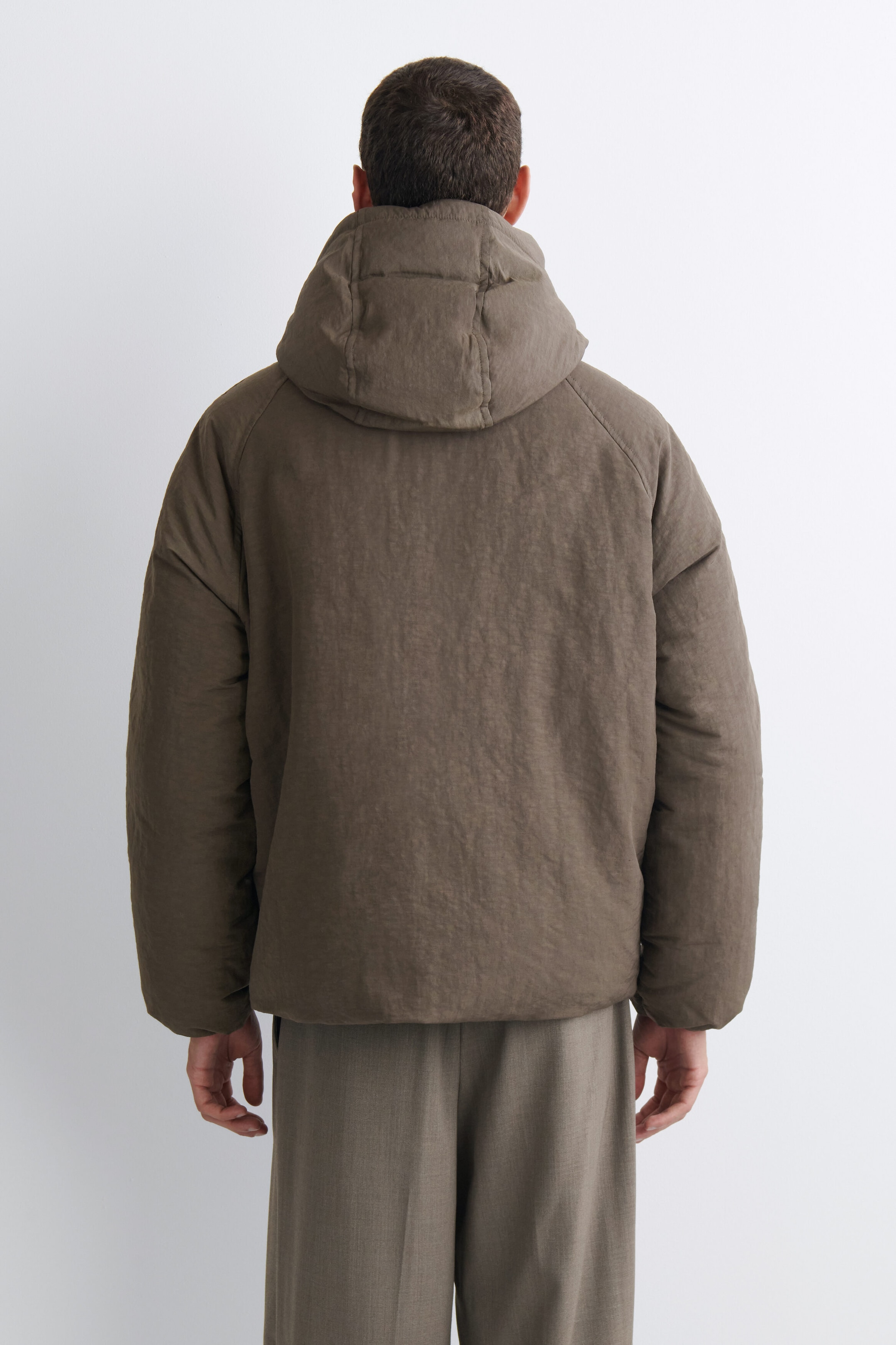 The oversized nylon-linen hooded down puffer jacket is shown from the back, featuring a large, puffy hood, raglan sleeves, and a textured taupe-brown finish. The jacket hangs loosely to create a boxy, voluminous silhouette, paired with matching trousers. The oversized nylon-linen hooded down puffer jacket is shown from the back, featuring a large, puffy hood, raglan sleeves, and a textured taupe-brown finish. The jacket hangs loosely to create a boxy, voluminous silhouette, paired with matching trousers.