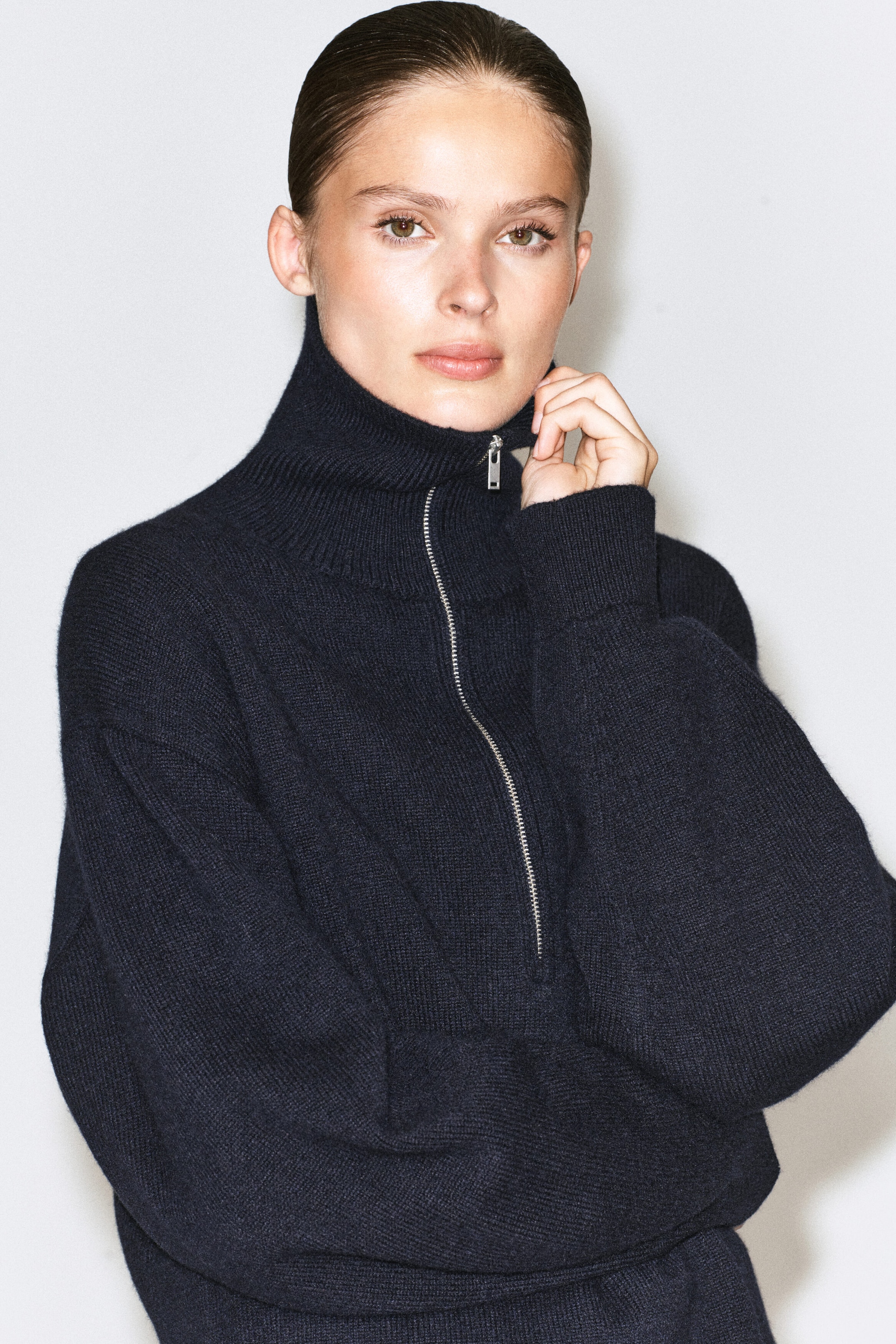 A dark navy knitted jumper featuring a ribbed high collar and a silver half-zip fastening is being worn by the model. The model is lightly touching the collar near the zip pull with one hand.