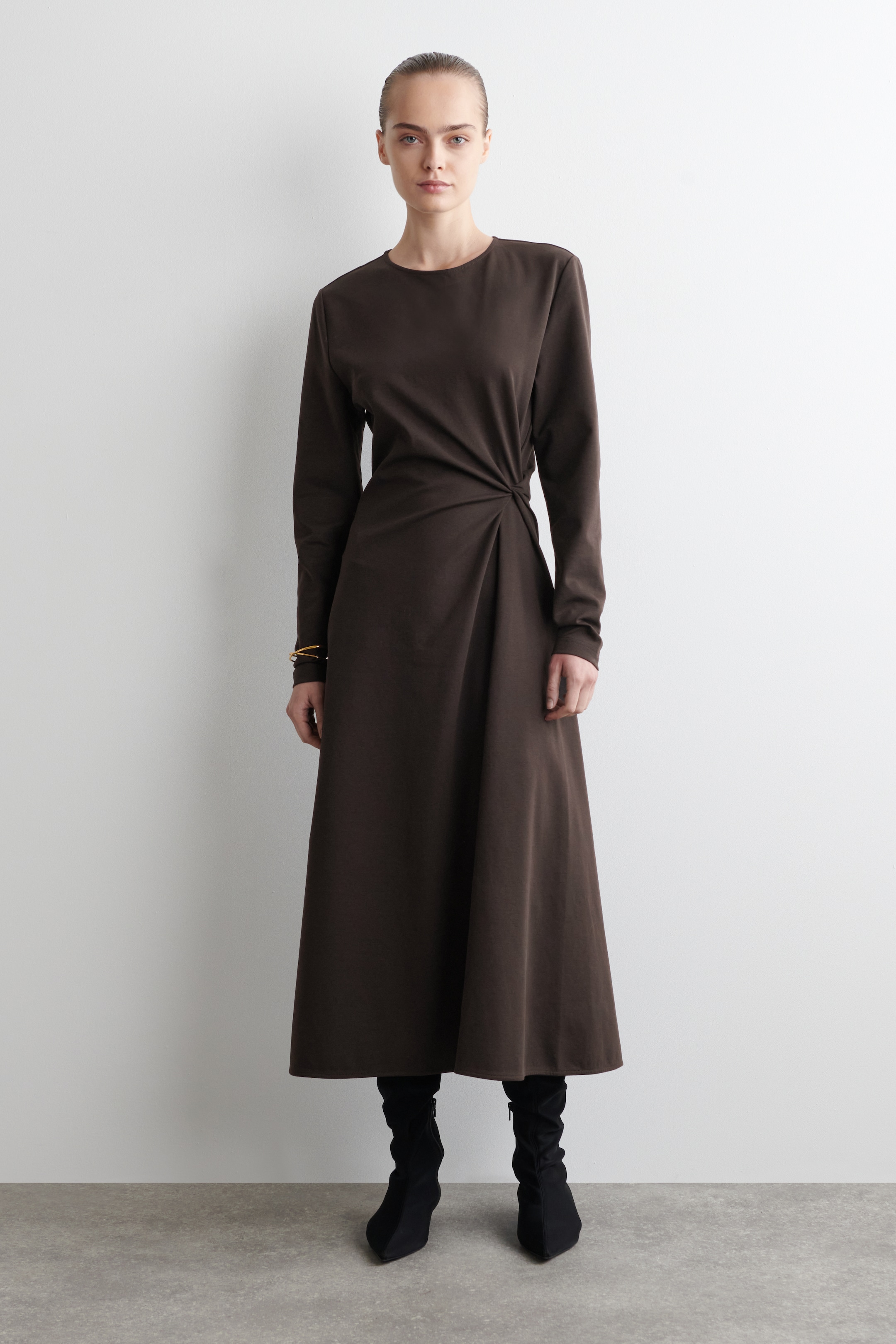 Agrandir l'image: Une robe midi à manches longues de couleur brun foncé est présentée de face, dotée d'un col rond et d'une jupe évasée. Un détail noué rassemble le tissu à la hanche gauche, créant des drapés, et elle est portée avec des bottes noires à bout pointu et un bracelet de couleur dorée au poignet droit.