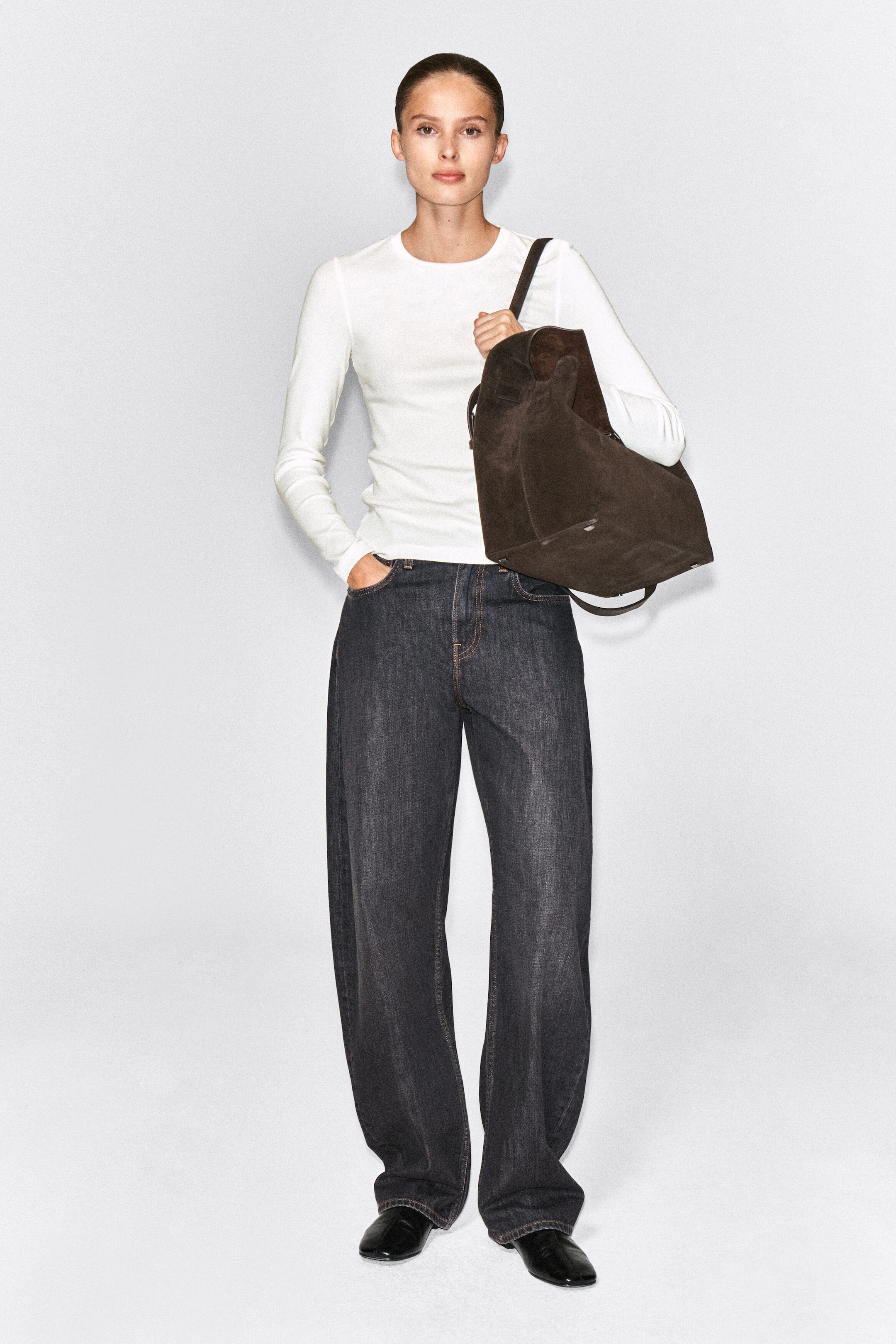 The model is wearing a slim, ribbed white cotton long-sleeve top with a crew neck and a close-fitting silhouette. It is styled with dark-wash wide-leg jeans, and the model is carrying a large brown suede shoulder bag. The model is wearing a slim, ribbed white cotton long-sleeve top with a crew neck and a close-fitting silhouette. It is styled with dark-wash wide-leg jeans, and the model is carrying a large brown suede shoulder bag.