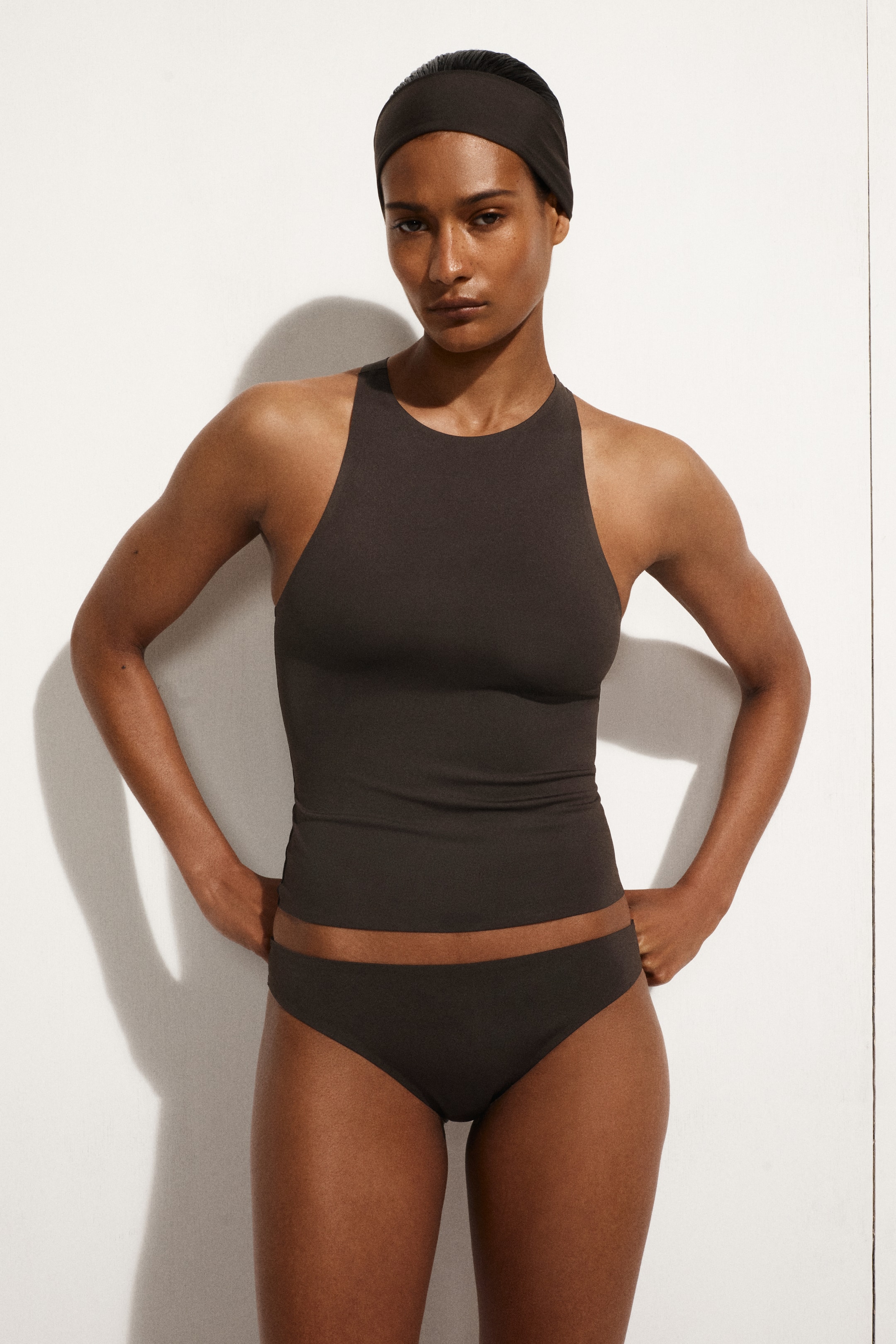 Größeres Bild ansehen: Ein dunkelbraunes Sportset wird präsentiert, bestehend aus einem Tanktop mit hohem Rundhalsausschnitt, passenden Slip-ähnlichen Unterteilen und einem breiten Stirnband. Alle Teile schmiegen sich eng an den Körper an.