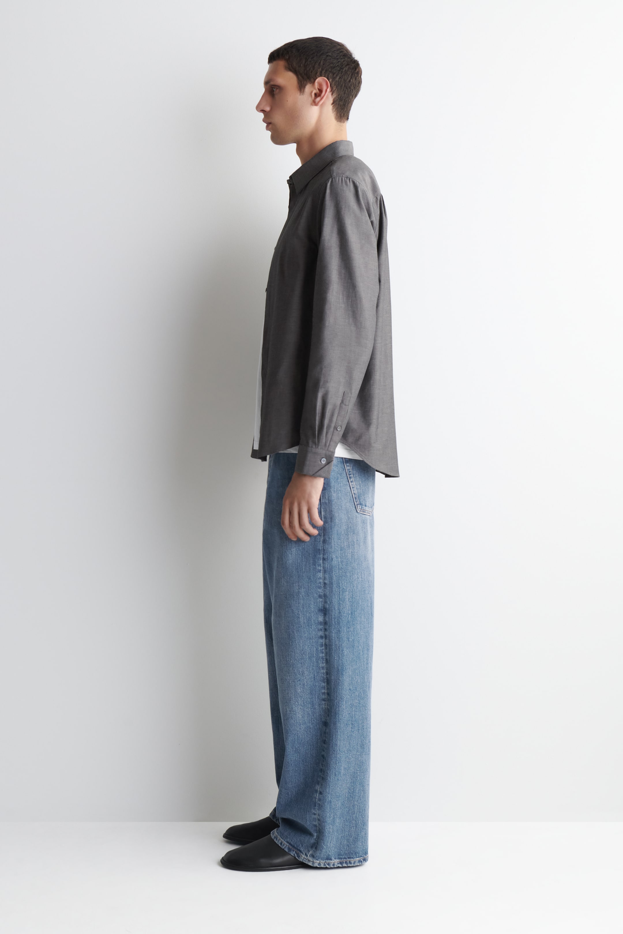 Visualizza l'immagine ingrandita: La camicia grigia è presentata in profilo laterale, con un colletto classico, maniche lunghe con polsini abbottonati e una vestibilità morbida, indossata non infilata sopra una t-shirt bianca.