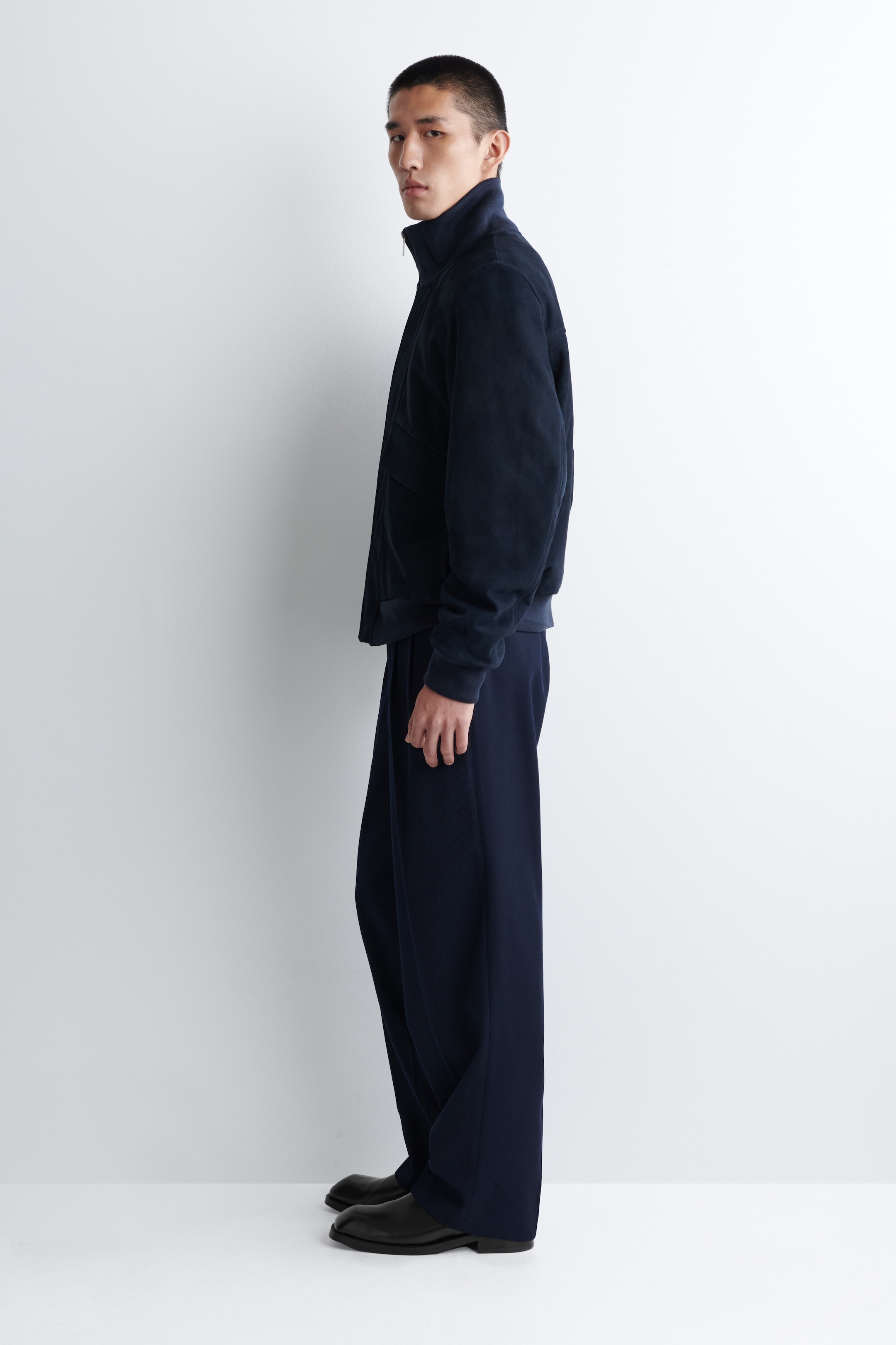 Visualizza immagine più grande: Una giacca blu scuro e pantaloni abbinati a gamba larga vengono presentati in un profilo laterale sinistro. La giacca presenta un colletto alto, una cerniera frontale a tutta lunghezza e polsini e orlo a fascia, mentre i pantaloni mostrano una vestibilità rilassata, completati da scarpe eleganti nere.