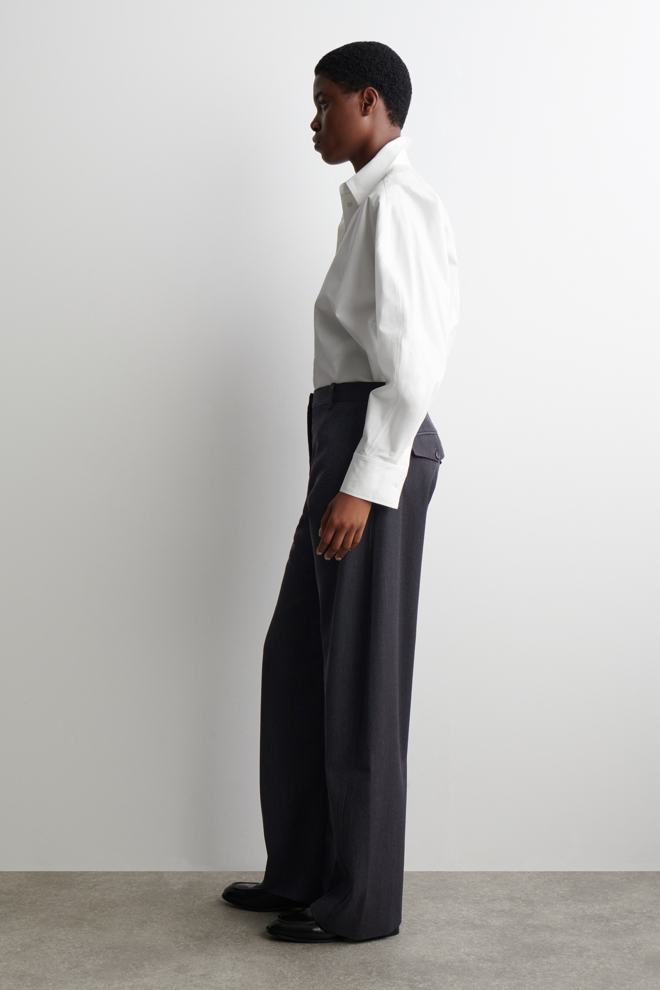 View larger image: A white collared shirt, featuring a structured collar and wide cuffs, is being worn tucked into dark wide-leg trousers. The side profile is showing the shirt's relaxed fit and the trousers' loose drape, which are falling over black flat shoes.