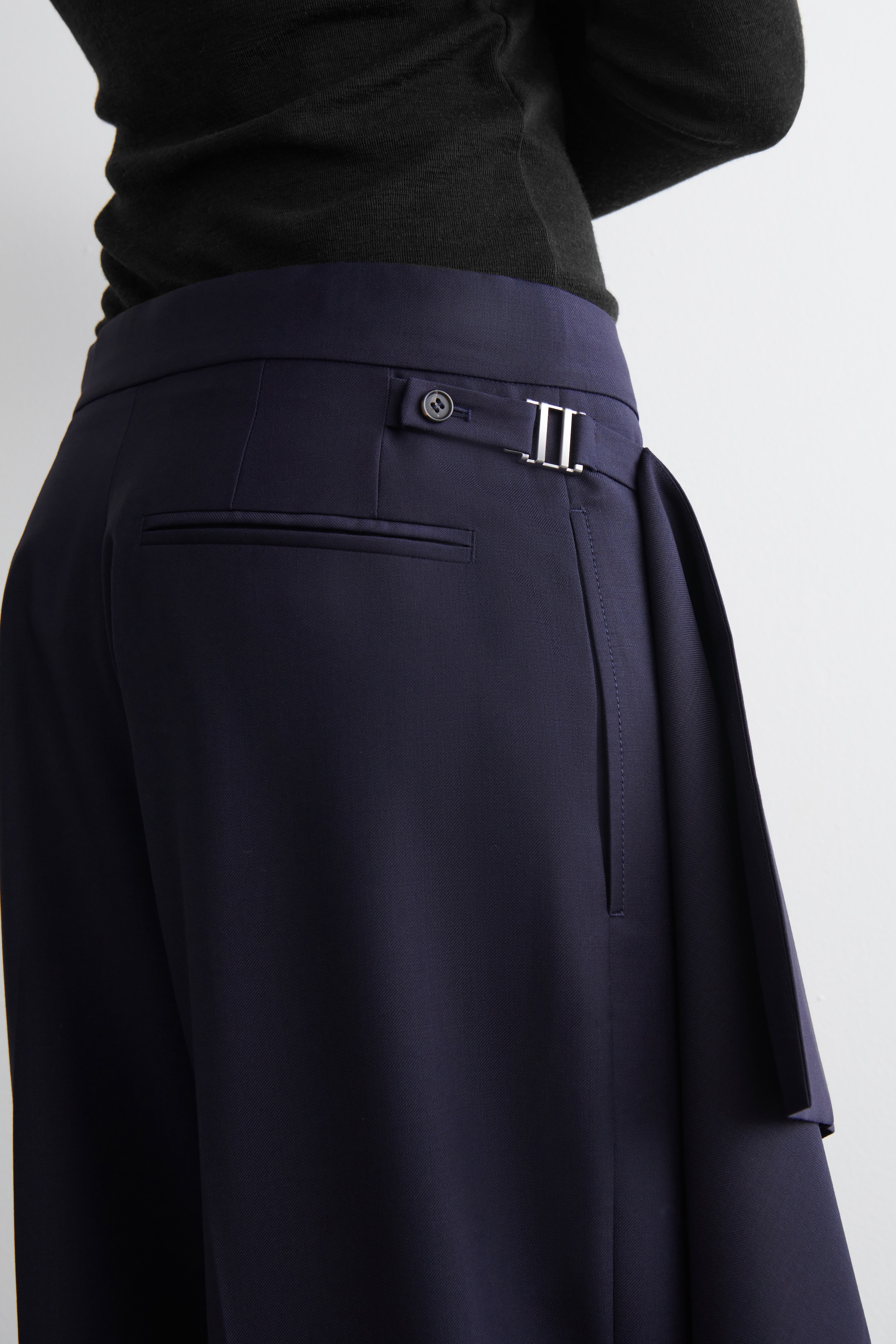 View larger image: Navy blue trousers are being presented from the back-right side, worn with a dark long-sleeved top. They feature a wide waistband with a buttoned side tab and a silver-toned double-bar buckle on the right, a jetted pocket on the back, and a structured fabric panel extending from the right side.