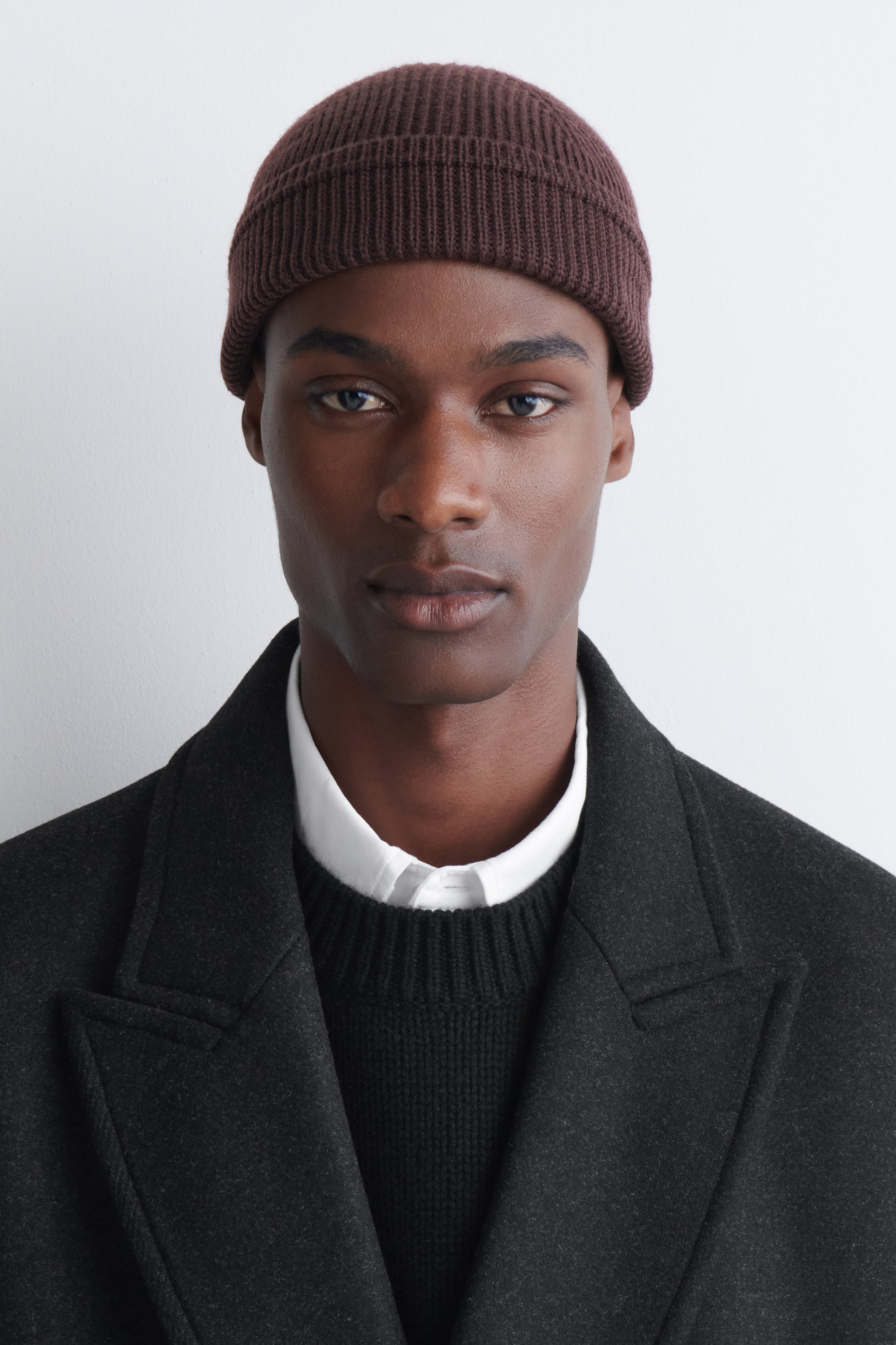 View larger image: A ribbed wool-cotton fisherman beanie in a dark brown shade is being worn by the model. The beanie features a classic ribbed texture with a folded cuff, styled with a dark wool overcoat layered over a black knitted jumper and a white collared shirt.