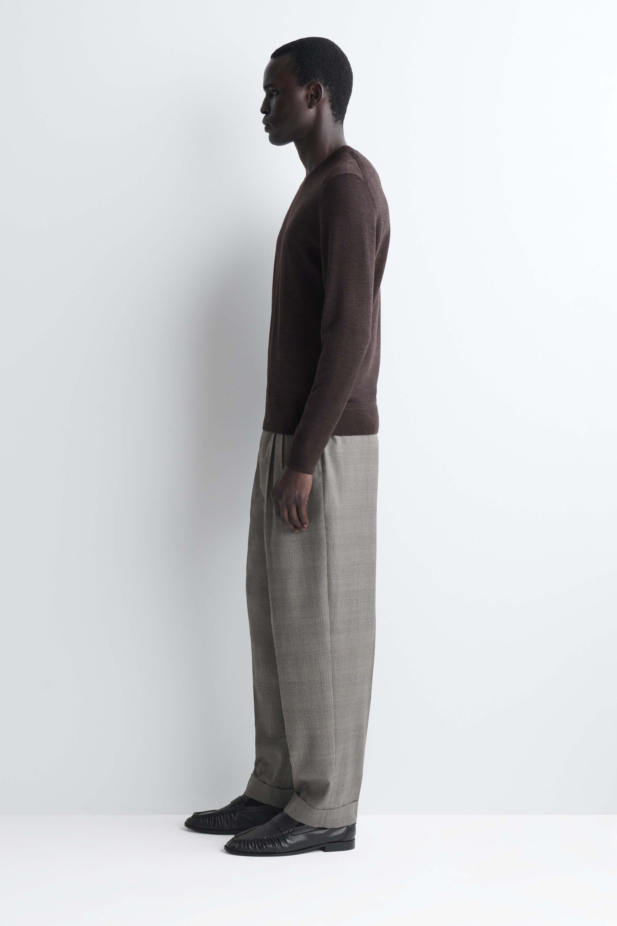 The slim merino wool crewneck sweater in dark brown is shown from a side profile facing left, highlighting its close-fitting silhouette on the torso and arms. The sweater features a fine-knit texture, ribbed cuffs, and a ribbed hem resting over the waistband of light gray trousers. The slim merino wool crewneck sweater in dark brown is shown from a side profile facing left, highlighting its close-fitting silhouette on the torso and arms. The sweater features a fine-knit texture, ribbed cuffs, and a ribbed hem resting over the waistband of light gray trousers.