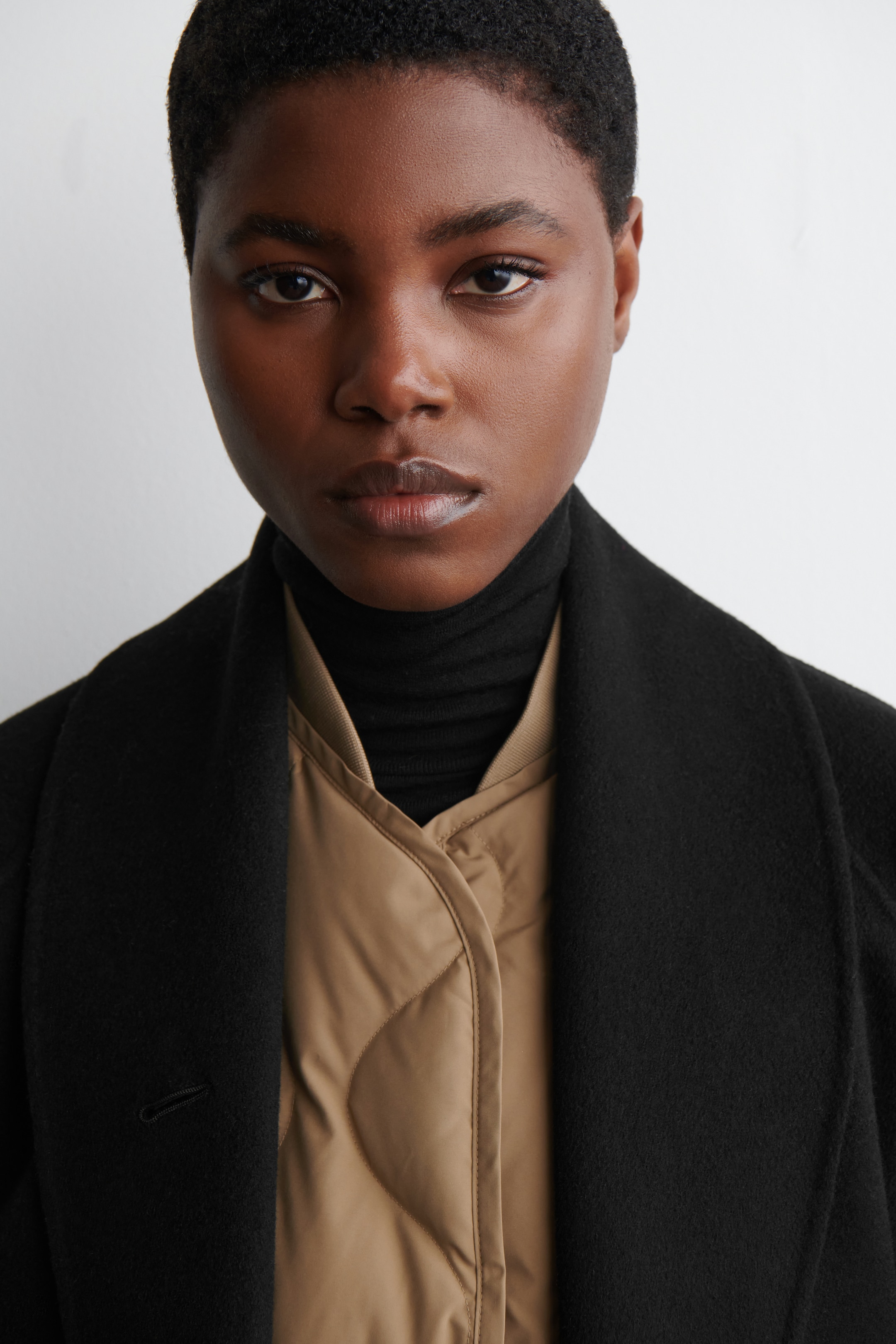 View larger image: A black coat with broad, open lapels is being worn from a front-facing angle, revealing a tan quilted jacket with diamond quilting and a black turtleneck sweater layered underneath. A single black button and buttonhole are visible on the coat's front.