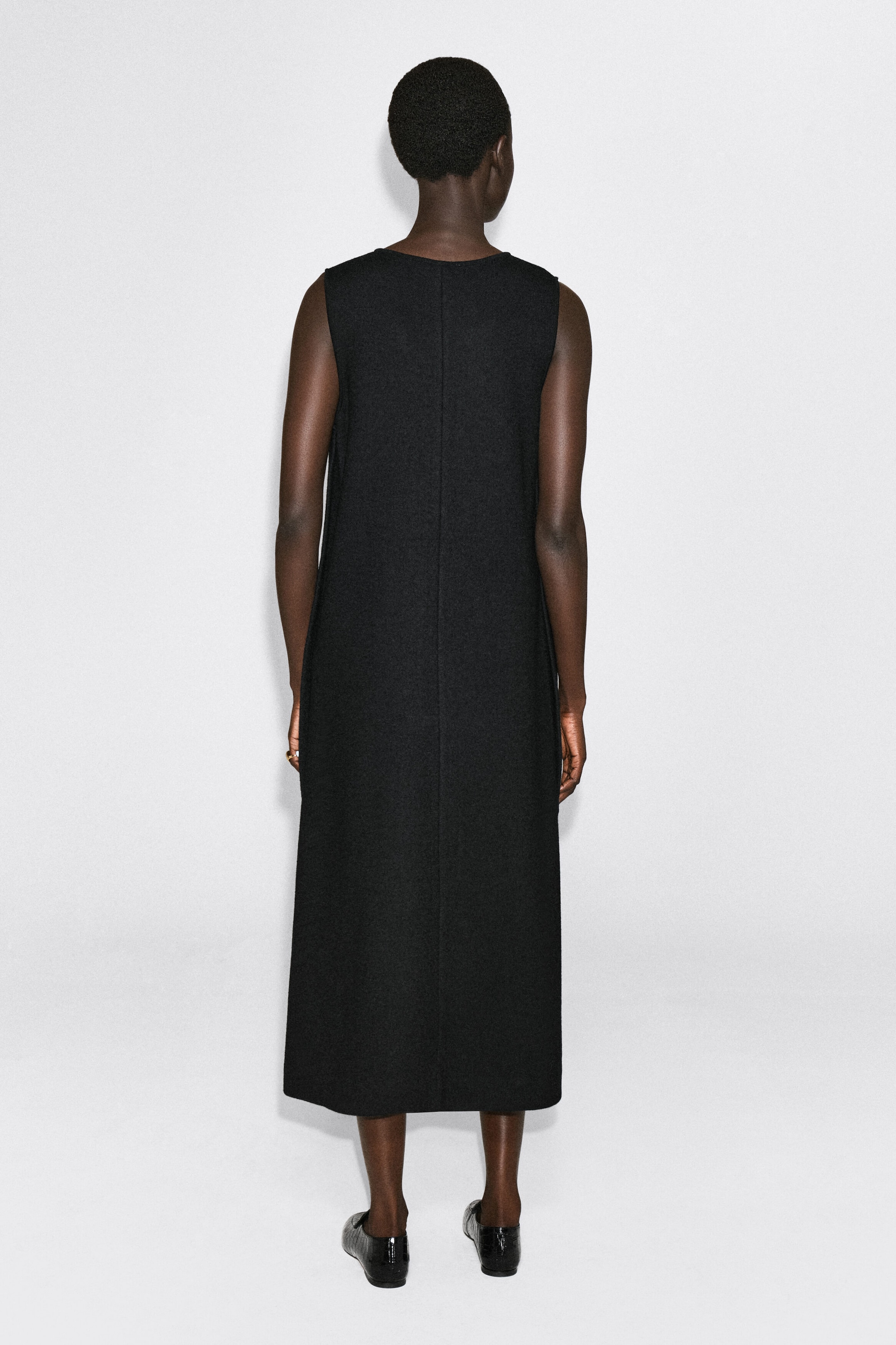 The boiled wool-blend V-neck midi dress drapes in a straight, sleeveless silhouette, shown from the back to highlight its structured fit and central vertical seam. The black textured dress falls to a midi-length hemline. The boiled wool-blend V-neck midi dress drapes in a straight, sleeveless silhouette, shown from the back to highlight its structured fit and central vertical seam. The black textured dress falls to a midi-length hemline.