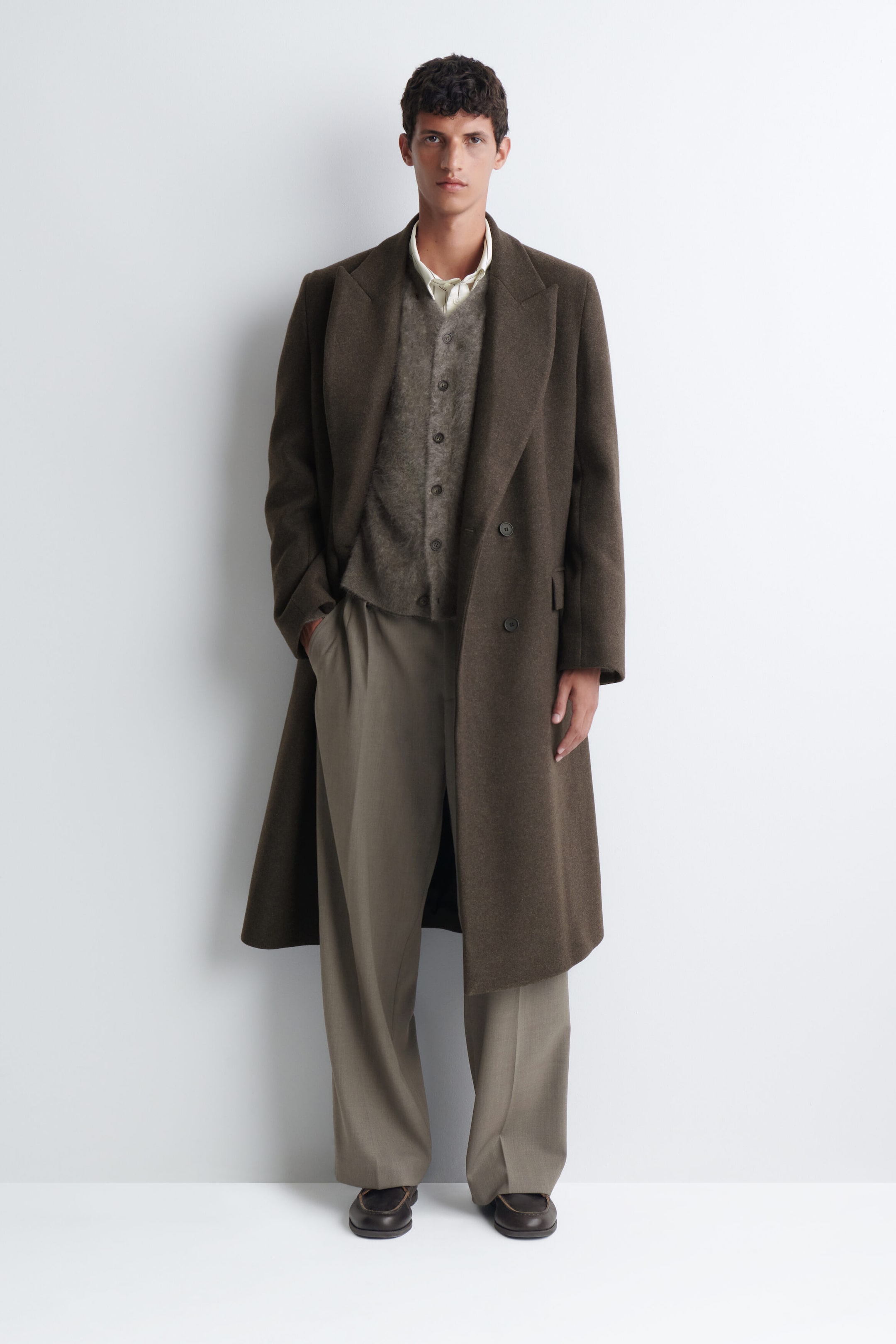 Un cardigan à col en V en cachemire brossé, dans une teinte gris-brun texturée, est porté par le mannequin, superposé à une chemise claire à col et sous un long manteau brun foncé. Le cardigan boutonné présente une texture douce et duveteuse et est stylisé avec un pantalon ample à jambe large. Un cardigan à col en V en cachemire brossé, dans une teinte gris-brun texturée, est porté par le mannequin, superposé à une chemise claire à col et sous un long manteau brun foncé. Le cardigan boutonné présente une texture douce et duveteuse et est stylisé avec un pantalon ample à jambe large.