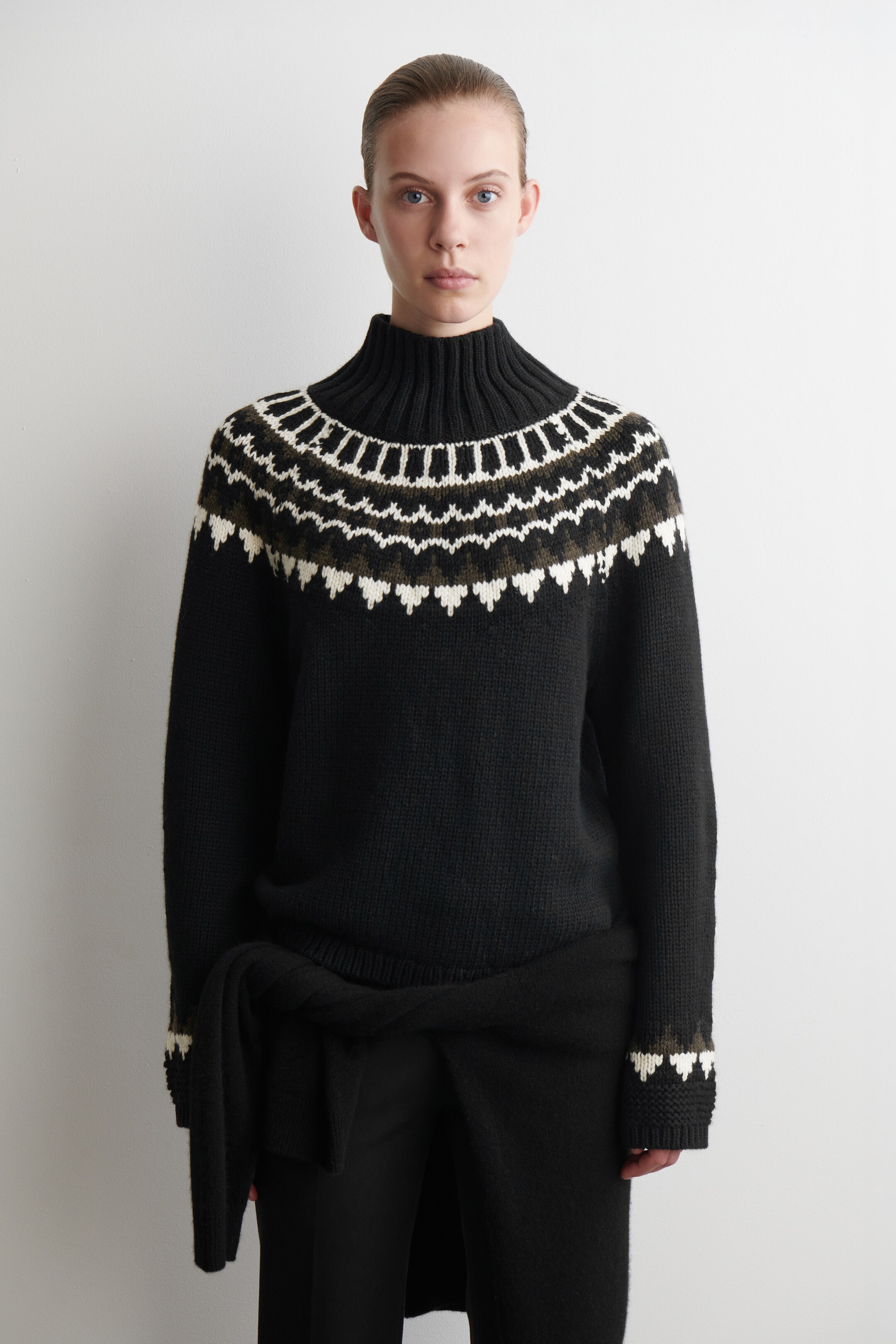 Ver imagem maior: Um camisola preta está a ser apresentada de frente, com uma gola alta canelada e um padrão fair isle em branco e verde-azeitona sobre o peito superior. As mangas compridas terminam em punhos canelados, mostrando também uma parte do padrão fair isle.