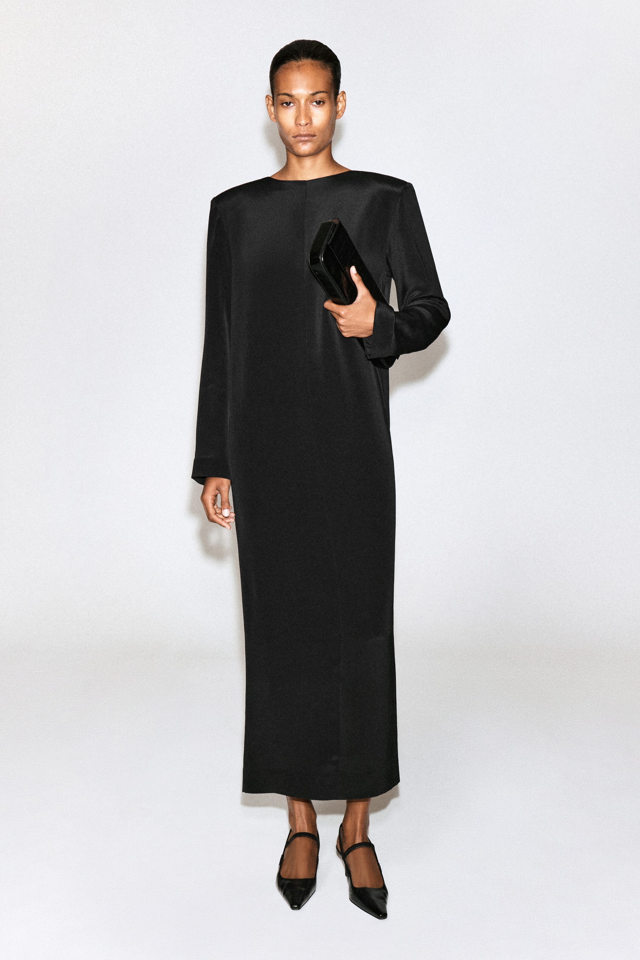 The model is wearing a black silk-crepe maxi dress with structured shoulders and long sleeves. She is holding a black clutch bag and wearing black pointed-toe shoes.