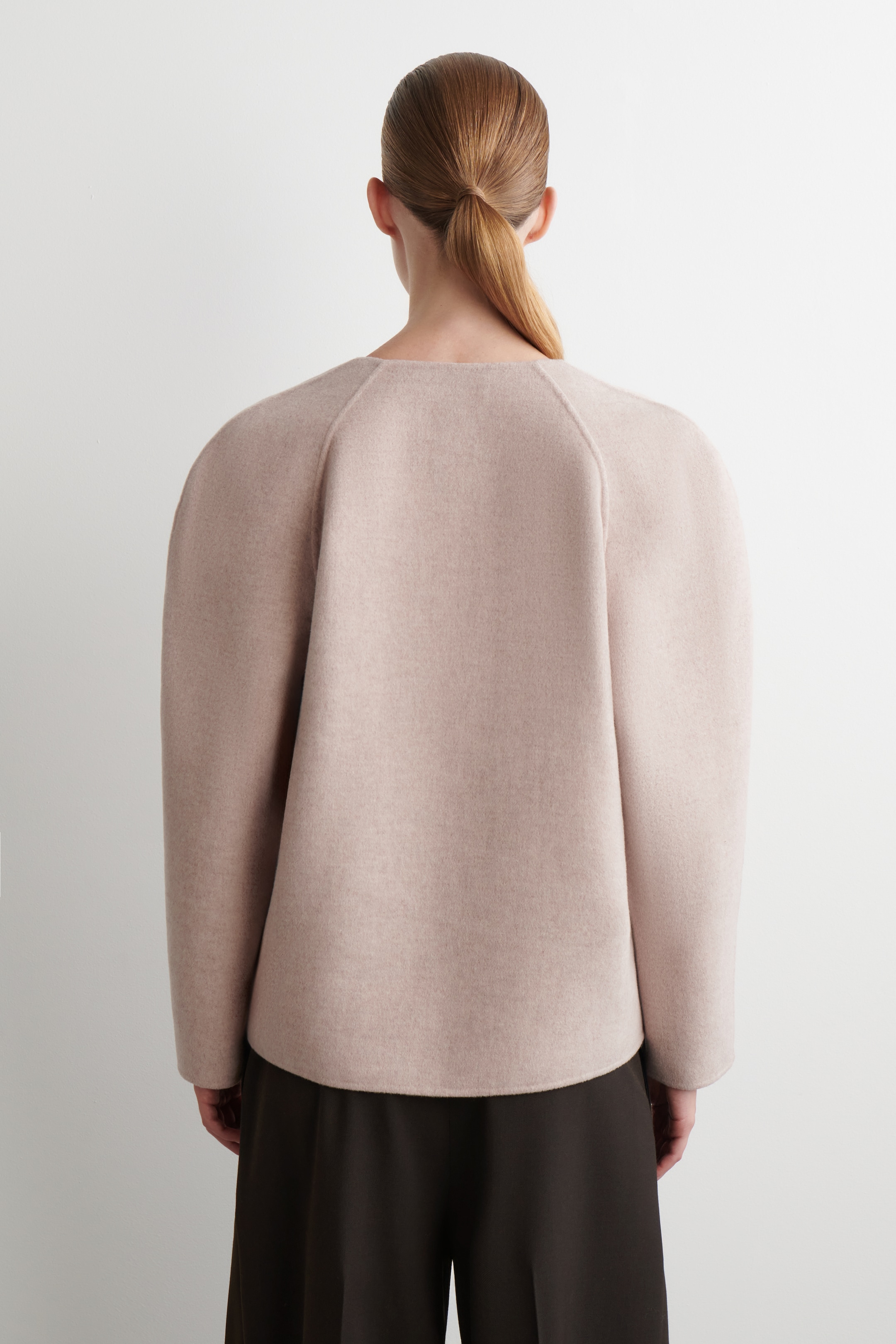 View larger image: A light pink, smooth-textured top is being viewed from the back. It is featuring wide, voluminous raglan sleeves, a rounded neckline, and a straight hemline that is sitting above dark trousers.