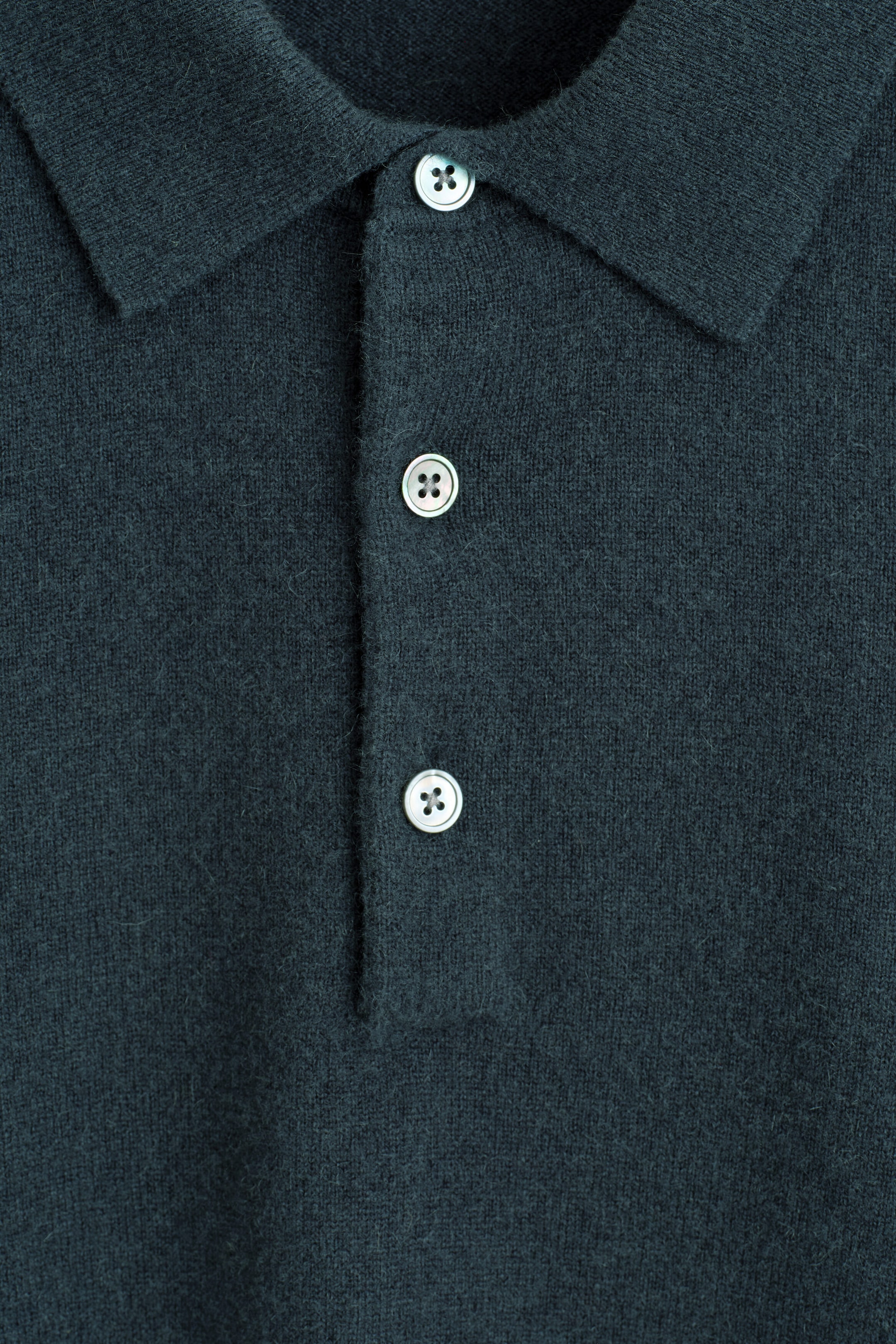 A cashmere polo sweater is shown from the front in a close-up view, highlighting its soft, dark teal-green knit texture. The garment features a classic polo collar and a central placket secured with three light-coloured buttons.