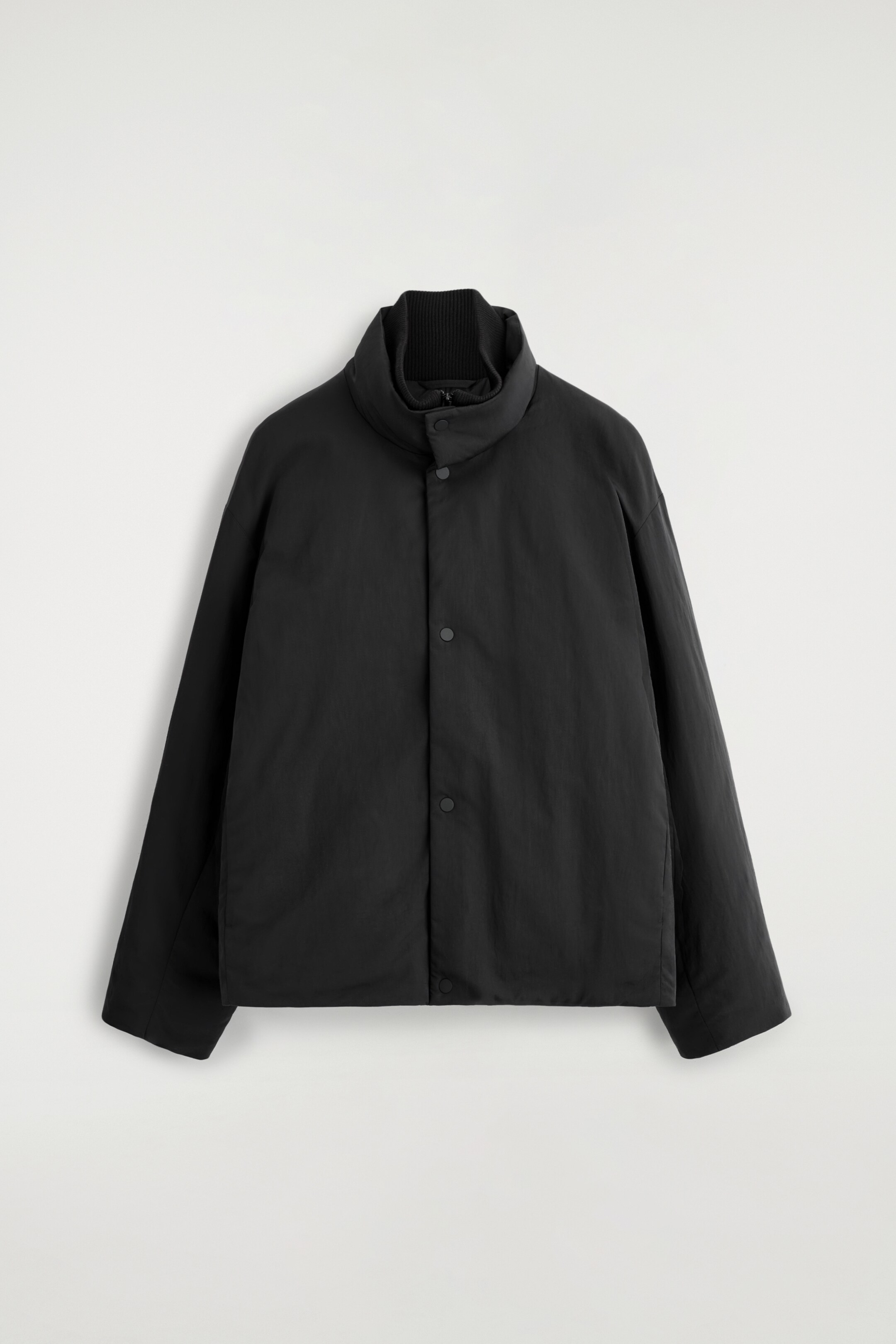 Größeres Bild ansehen: Eine schwarze wattierte Jacke wird von vorne präsentiert und zeigt eine entspannte, kastige Passform. Sie verfügt über einen hohen Trichterkragen mit einem sichtbaren gerippten Innenkragen, und der Verschluss vorne wird durch eine Reihe schwarzer Druckknöpfe gesichert.