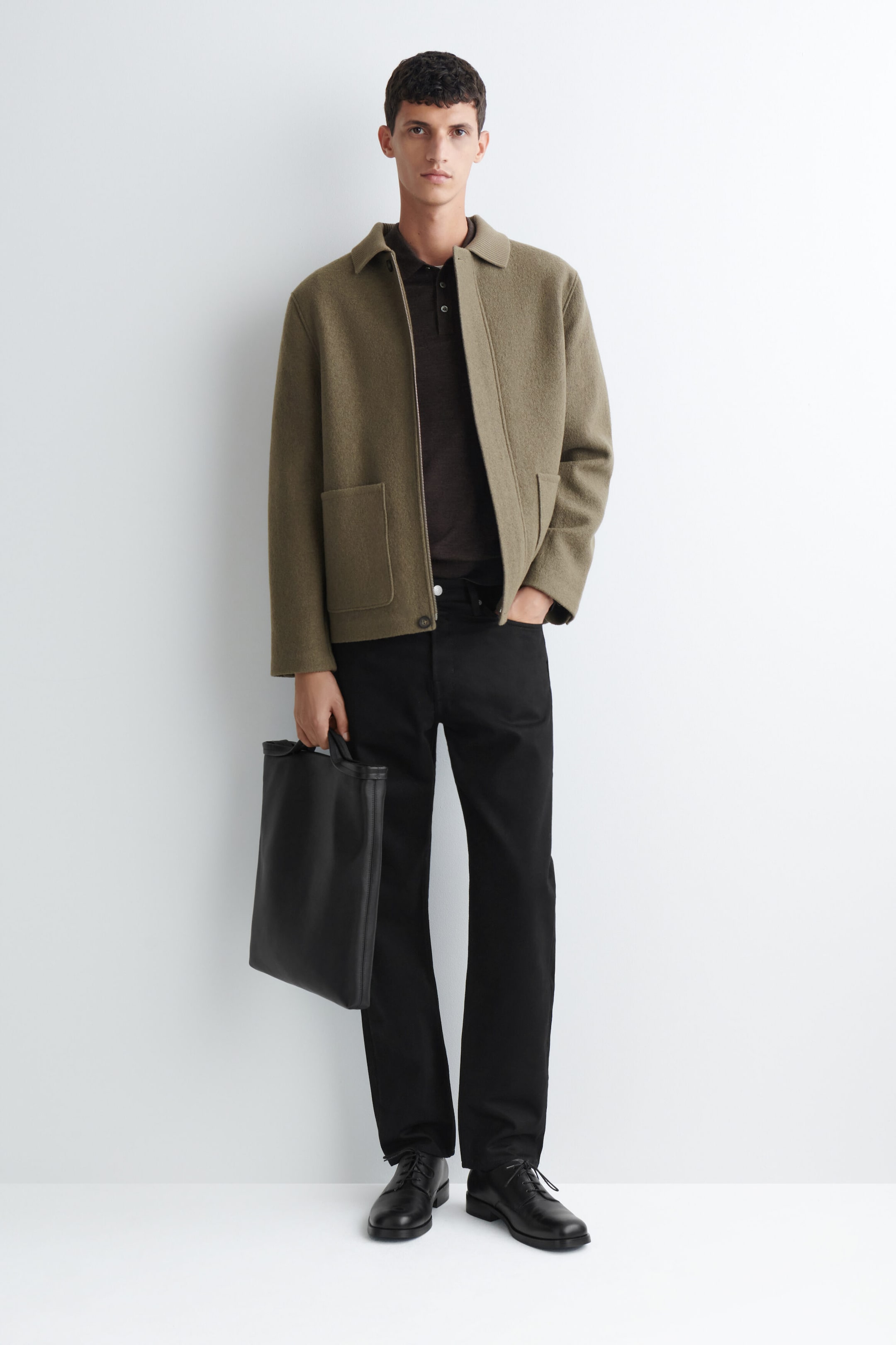 A ribbed-collar boiled-wool chore jacket in olive green is worn open by the model, highlighting its textured finish, classic collar, and front patch pockets. The jacket is layered over a dark polo shirt and styled with black trousers, while the model carries a black leather bag. A ribbed-collar boiled-wool chore jacket in olive green is worn open by the model, highlighting its textured finish, classic collar, and front patch pockets. The jacket is layered over a dark polo shirt and styled with black trousers, while the model carries a black leather bag.