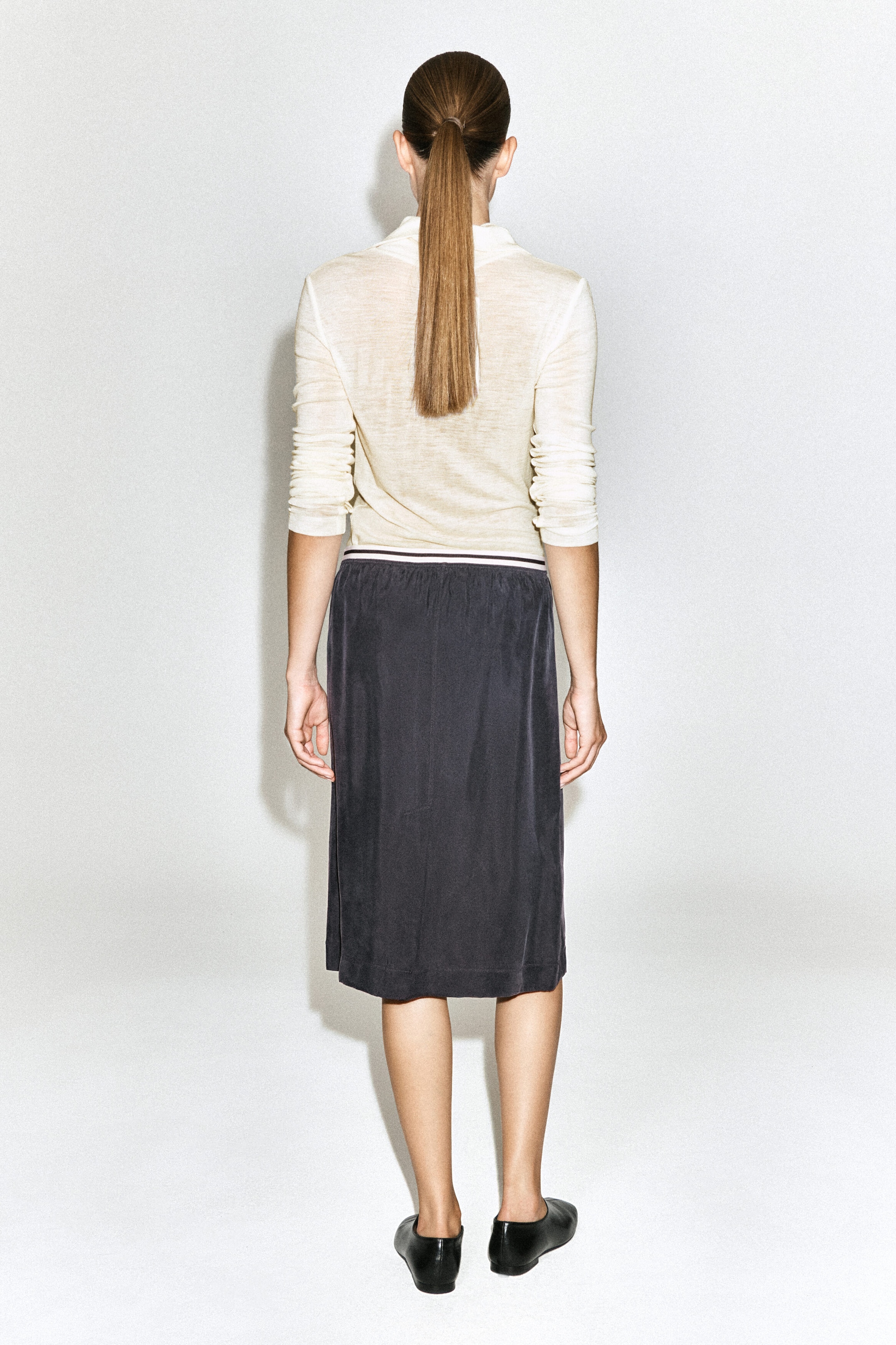 A dark gray contrast-panel pencil skirt is being worn by the model, featuring a straight silhouette and a contrasting striped waistband. The skirt is styled with a cream long-sleeved top tucked into the waistband, shown from the back. A dark gray contrast-panel pencil skirt is being worn by the model, featuring a straight silhouette and a contrasting striped waistband. The skirt is styled with a cream long-sleeved top tucked into the waistband, shown from the back.