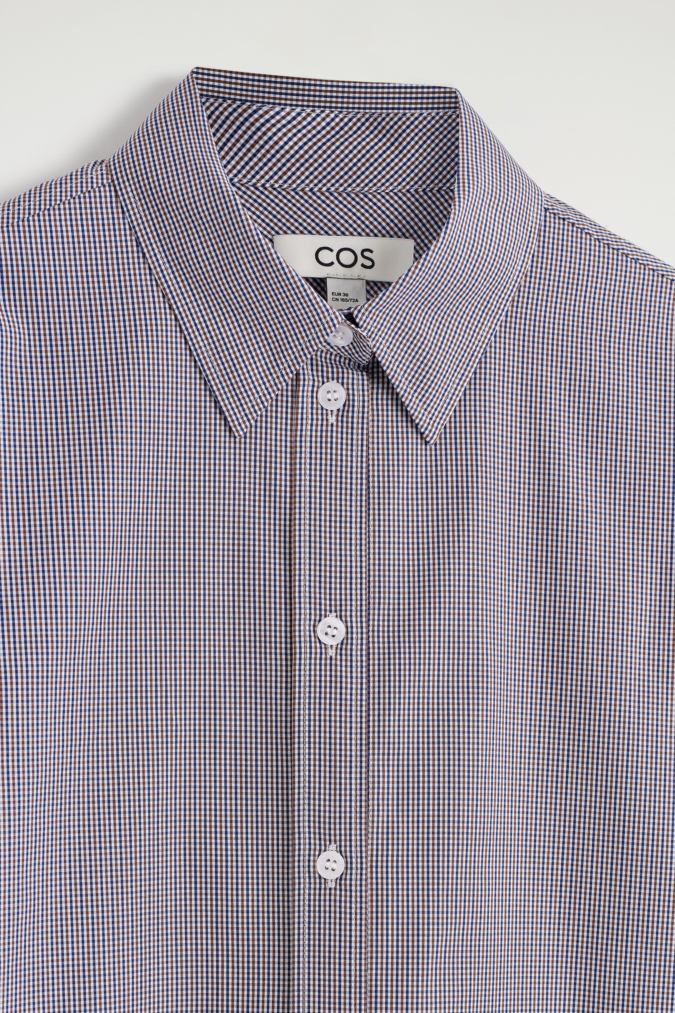 Visualizza immagine più grande: Una camicia mostra il suo FRONTE, caratterizzato da un motivo a quadri gingham blu scuro e marrone. È progettata con un colletto a punta e una chiusura frontale con bottoni bianchi.
