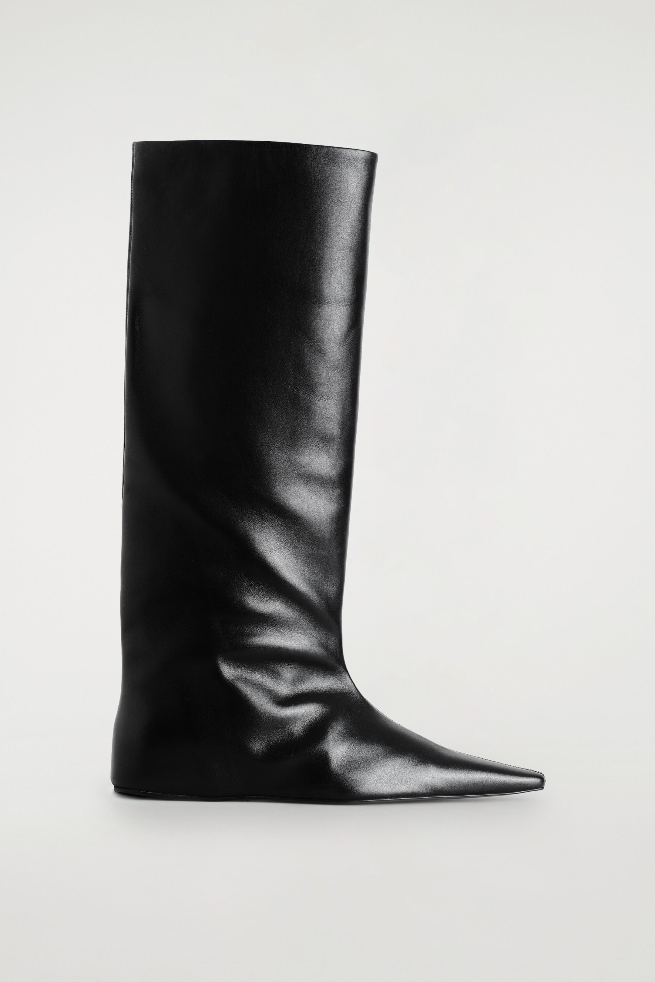 A black leather knee-high boot stands upright and faces right, showcasing an elongated pointed toe and a flat sole. The boot features a wide, structured shaft with soft creasing around the ankle. A black leather knee-high boot stands upright and faces right, showcasing an elongated pointed toe and a flat sole. The boot features a wide, structured shaft with soft creasing around the ankle.