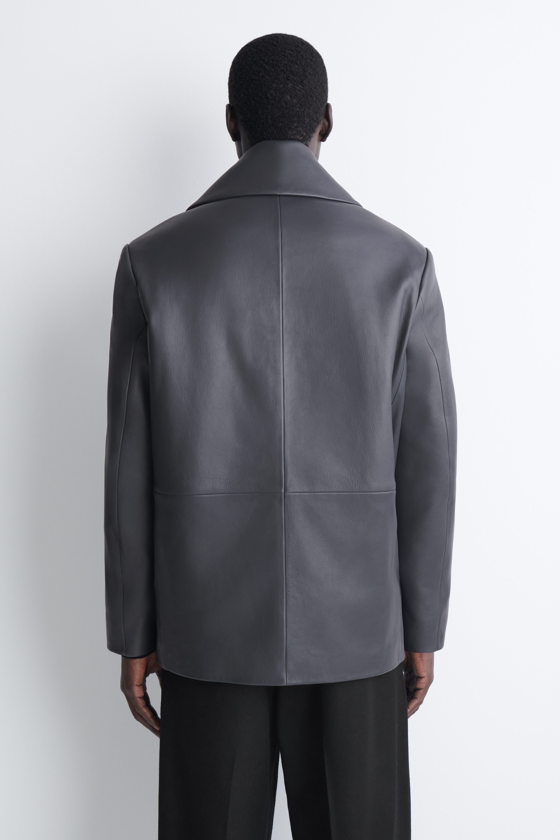 Eine dunkelgraue Lederjacke ist von hinten zu sehen und zeigt ein strukturiertes Design mit einer vertikalen Mittelnaht, einer horizontalen Naht im mittleren Rückenbereich und einem breiten Kragen. Die Jacke wird mit einer schwarzen Hose getragen, wodurch ihr gerade geschnittener Saum und die lockere Passform betont werden. Eine dunkelgraue Lederjacke ist von hinten zu sehen und zeigt ein strukturiertes Design mit einer vertikalen Mittelnaht, einer horizontalen Naht im mittleren Rückenbereich und einem breiten Kragen. Die Jacke wird mit einer schwarzen Hose getragen, wodurch ihr gerade geschnittener Saum und die lockere Passform betont werden.