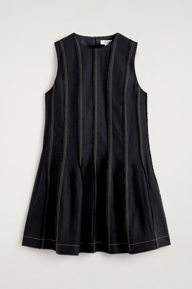 Une robe noire sans manches de coupe évasée, dotée d'un col rond et de plusieurs panneaux verticaux avec des coutures claires et des bords effilochés sur le devant, avec une poche latérale discrète visible sur la hanche droite. - 1