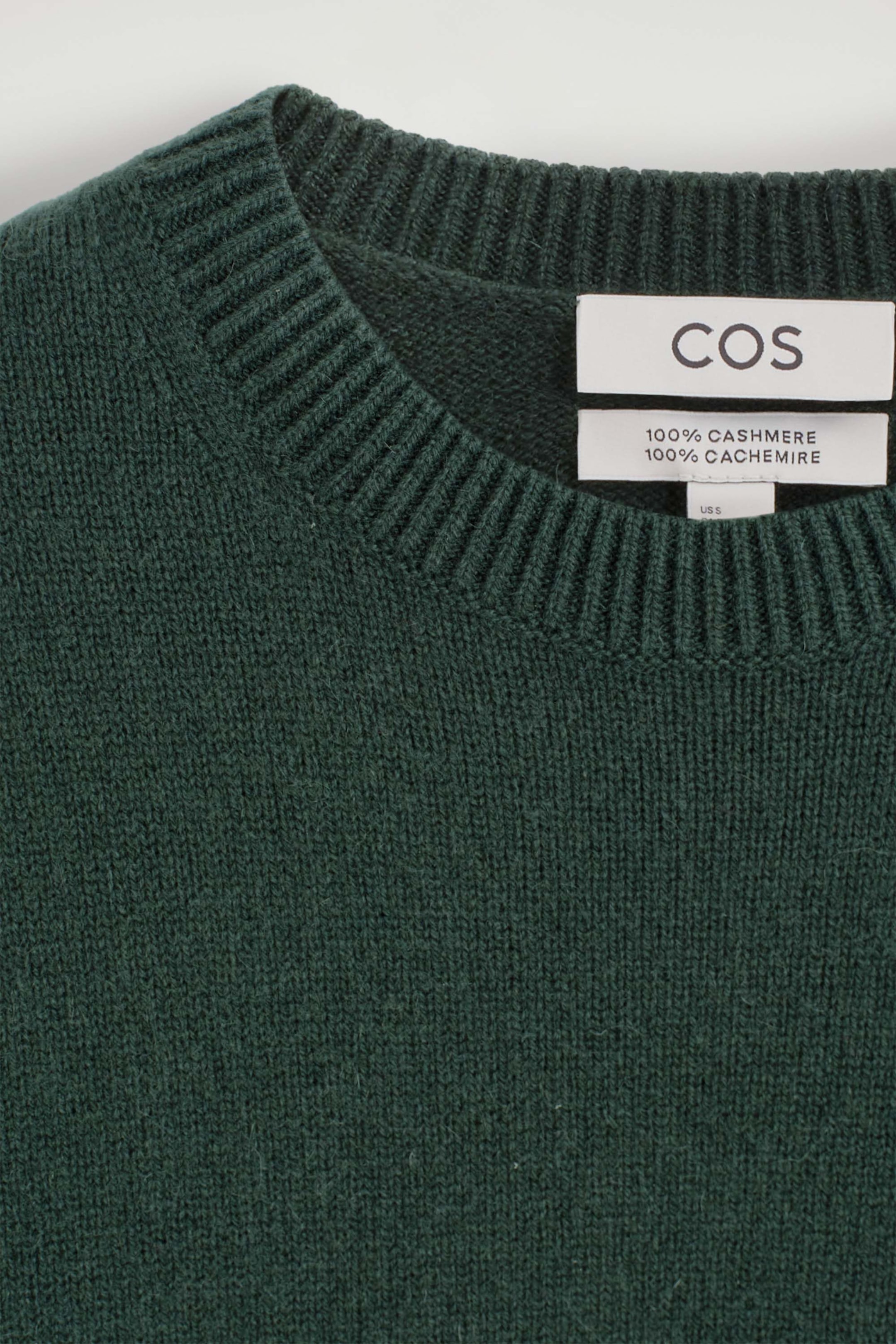 A close-up front view of a dark green cashmere crewneck sweater showcasing its finely knitted texture and ribbed neckline. The inner collar displays a white brand label alongside a material composition label. A close-up front view of a dark green cashmere crewneck sweater showcasing its finely knitted texture and ribbed neckline. The inner collar displays a white brand label alongside a material composition label.