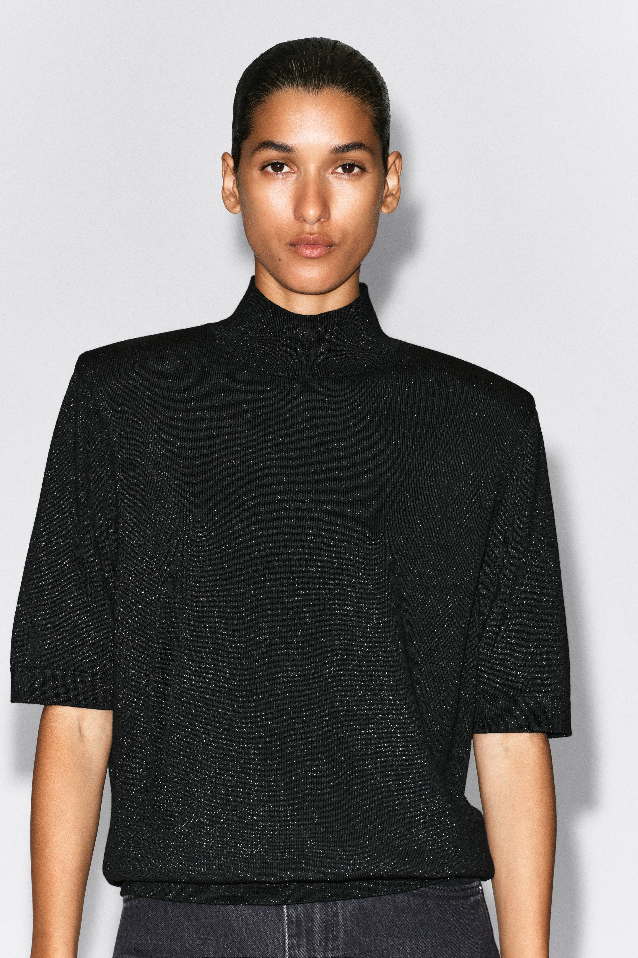 View larger image: A black knitted mock-neck top with a subtle metallic shimmer is shown from the front on a model. The top is designed with structured, sculpted shoulders, elbow-length sleeves with ribbed cuffs, and a matching ribbed mock neck.