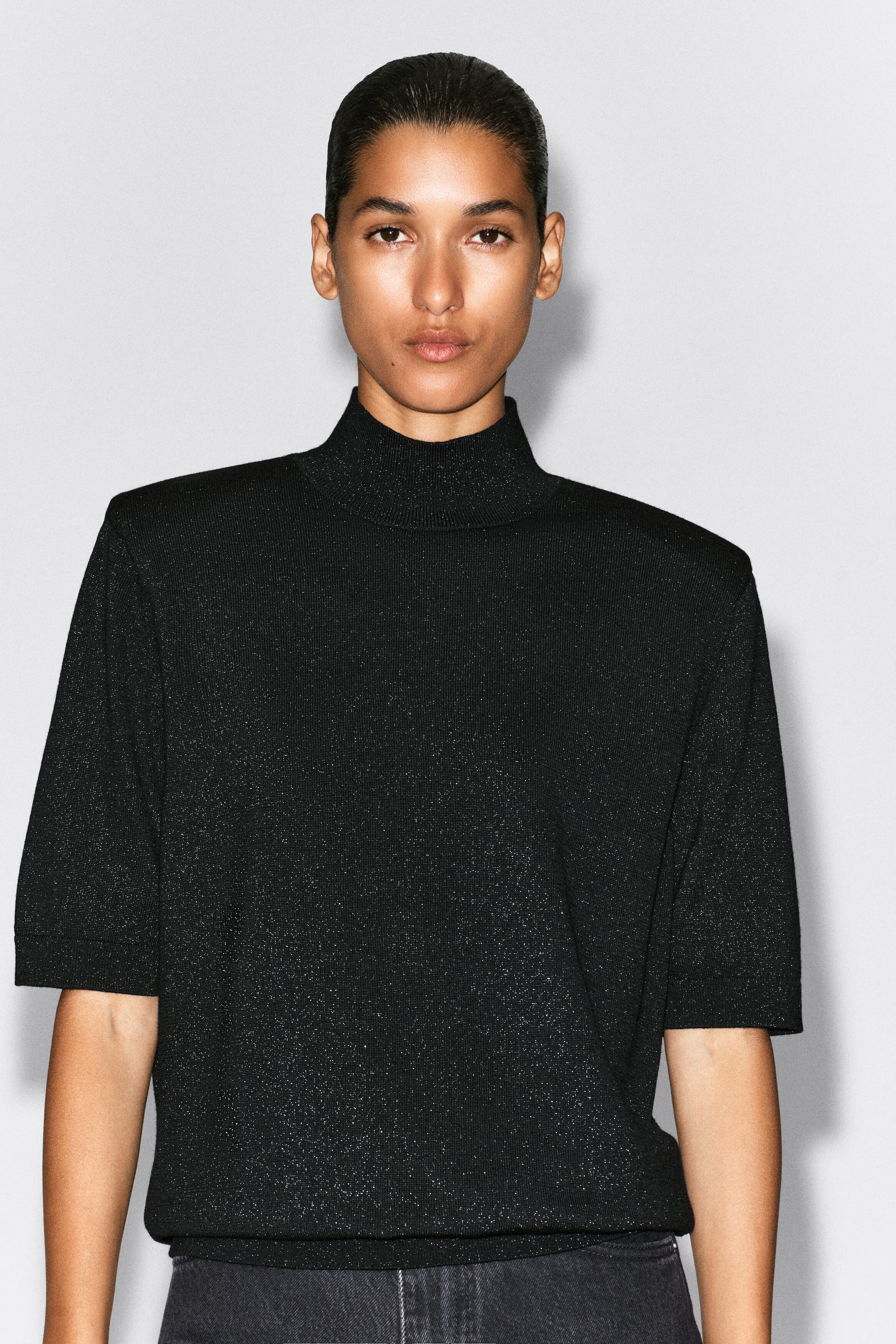 A black knit mock-neck top with a subtle metallic sheen is shown from the front on a model. The top features structured, sculpted shoulders, elbow-length sleeves with ribbed cuffs, and a matching ribbed mock neck. A black knit mock-neck top with a subtle metallic sheen is shown from the front on a model. The top features structured, sculpted shoulders, elbow-length sleeves with ribbed cuffs, and a matching ribbed mock neck.