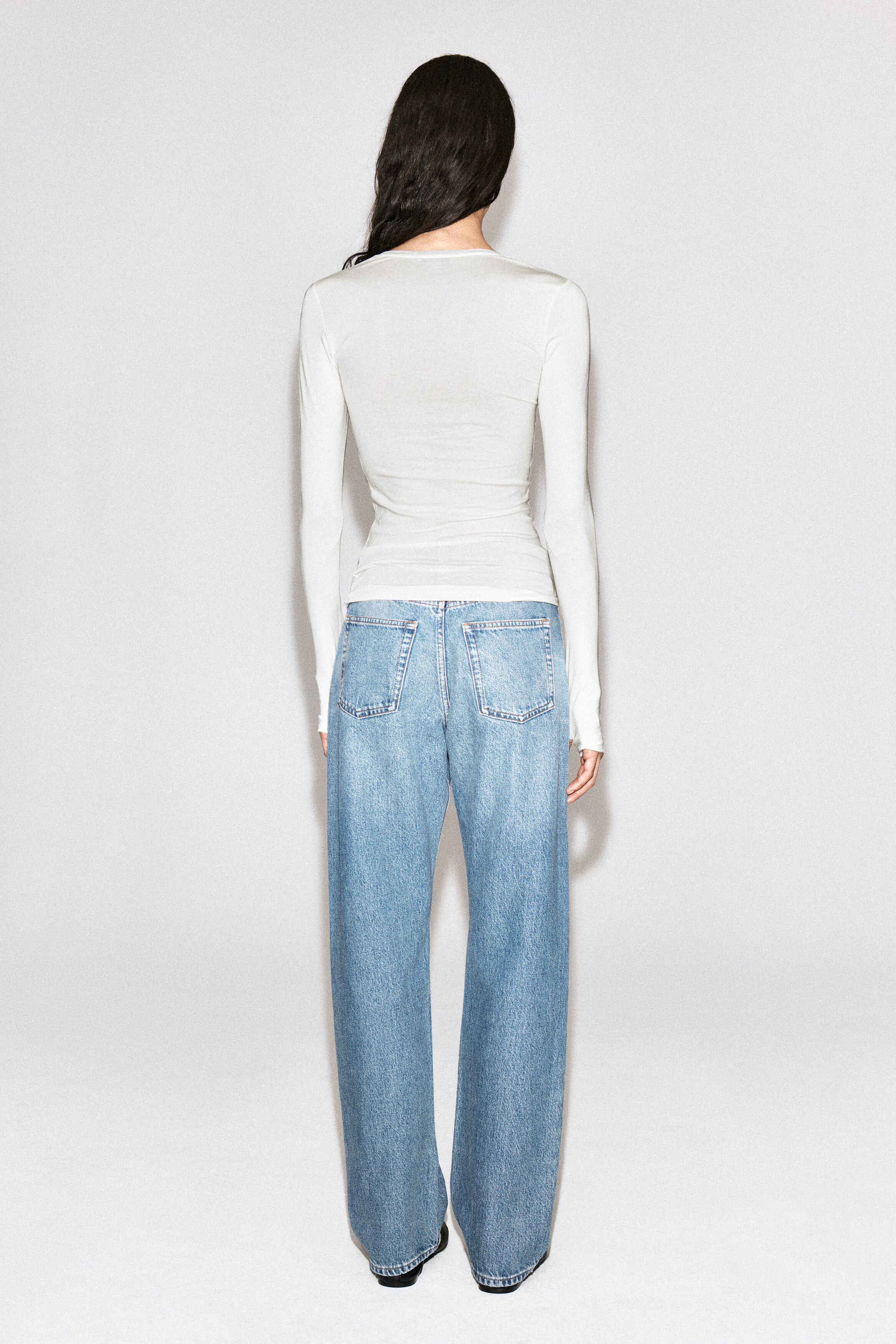 The model is shown from behind wearing light blue, wide-leg denim jeans. The jeans have a relaxed fit with two classic back patch pockets and are styled with a fitted white long-sleeve top.