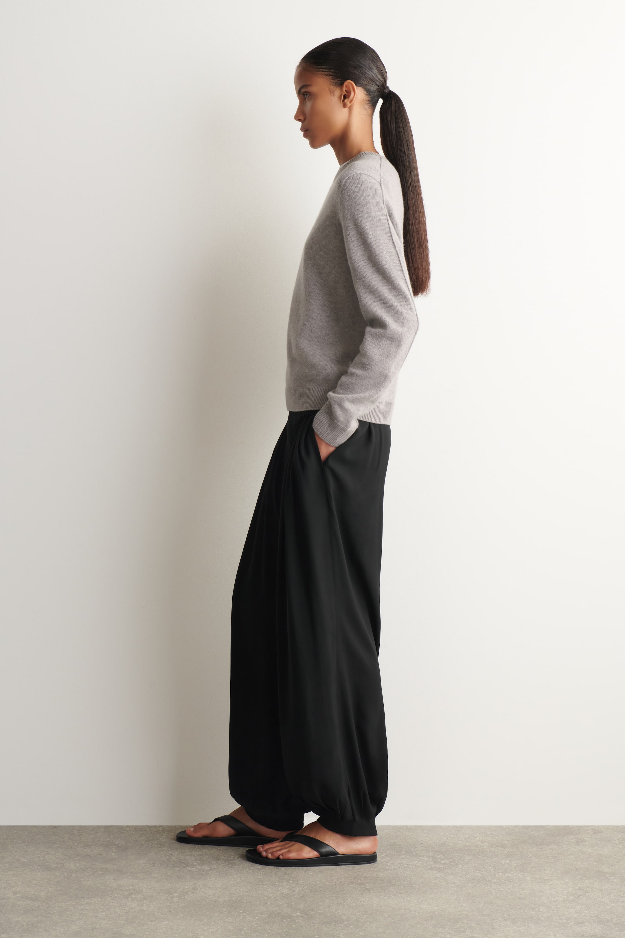 Agrandir l'image: Un pull gris à col rond avec des manches longues est porté, orienté vers la gauche. Il est associé à un pantalon noir à jambes larges avec une coupe fluide et des chevilles froncées, tombant sur des sandales plates noires.