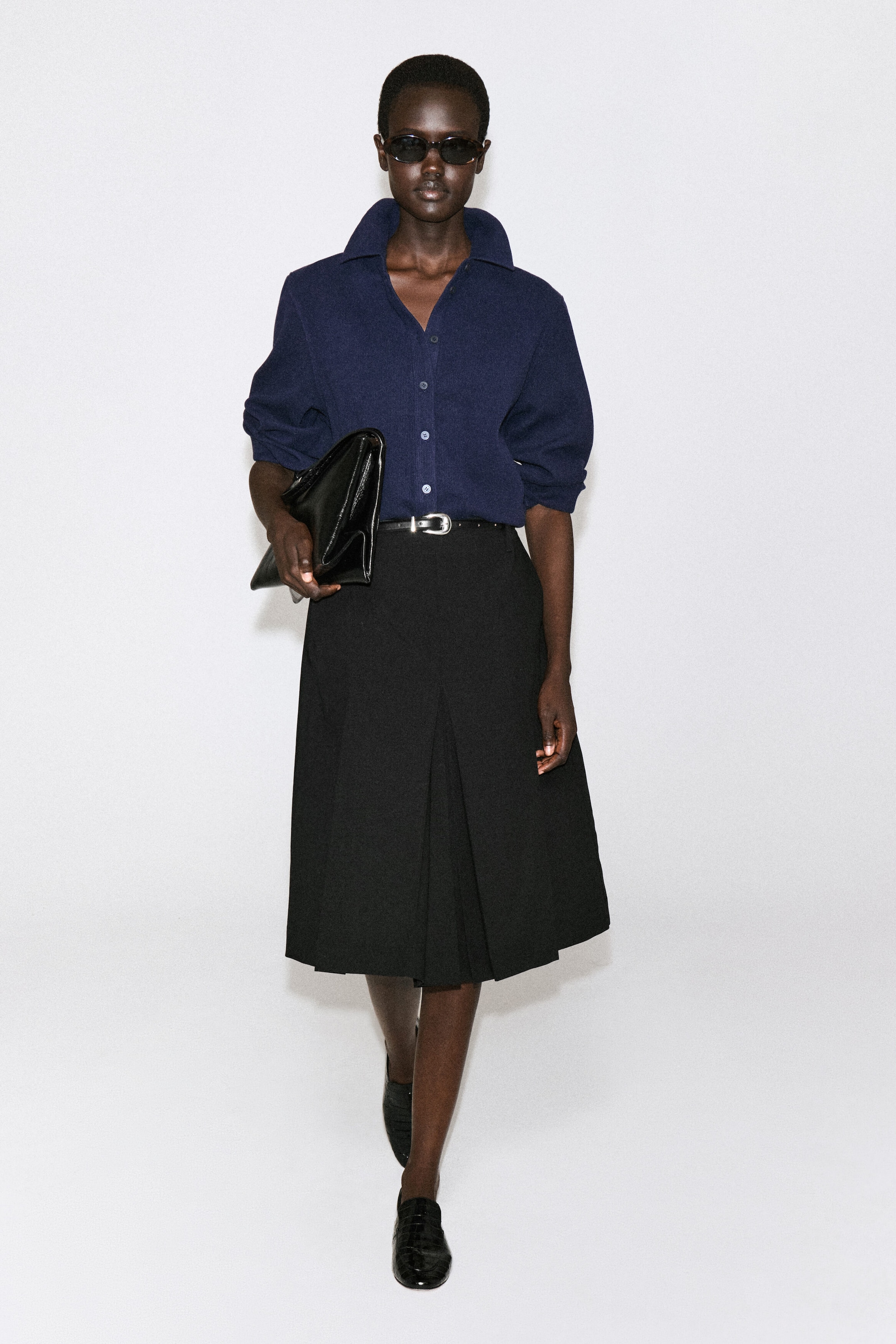 A navy blue corduroy-jersey shirt is shown from the front, featuring a relaxed fit silhouette with a pointed collar and button-front closure. The shirt is tucked into a high-waisted skirt, with the long sleeves styled pushed up to the elbows. A navy blue corduroy-jersey shirt is shown from the front, featuring a relaxed fit silhouette with a pointed collar and button-front closure. The shirt is tucked into a high-waisted skirt, with the long sleeves styled pushed up to the elbows.