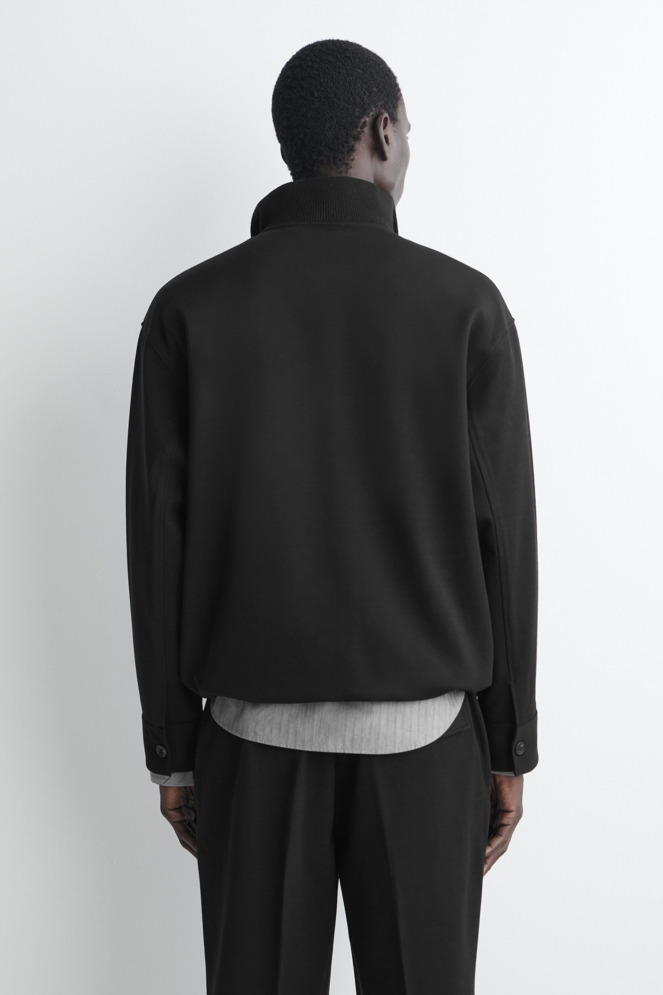 A black jacket is shown from the back, featuring a high ribbed stand collar, dropped shoulders, and long sleeves with button-fastened cuffs. The jacket’s straight hem is layered over a light gray shirt visible underneath, paired with matching black trousers.