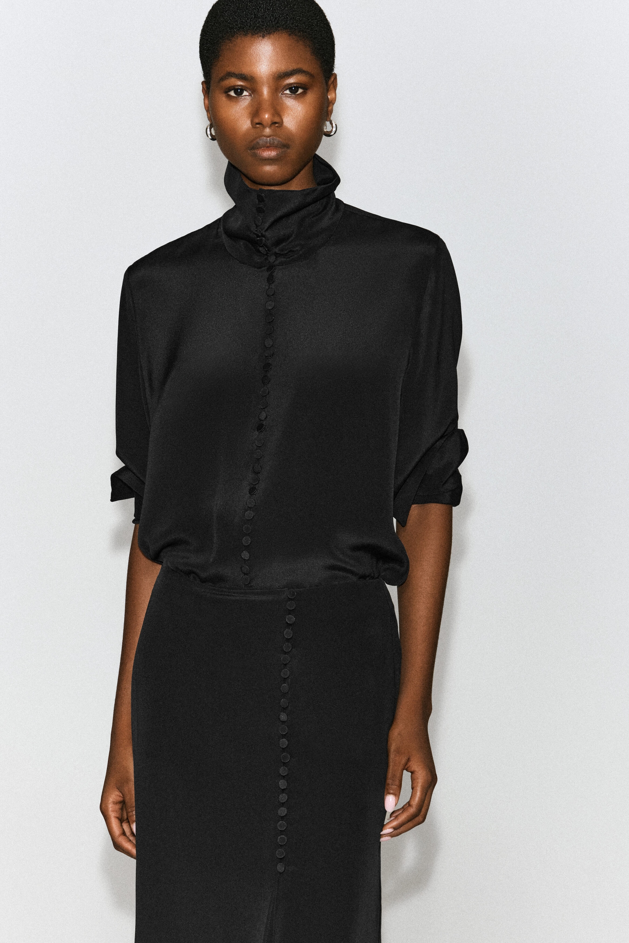 A black satin blouse featuring a high draped collar and a central row of small fabric-covered buttons is being worn by the model. The blouse has its sleeves rolled up to the elbows and is tucked into a matching black skirt with a coordinating button-front detail.