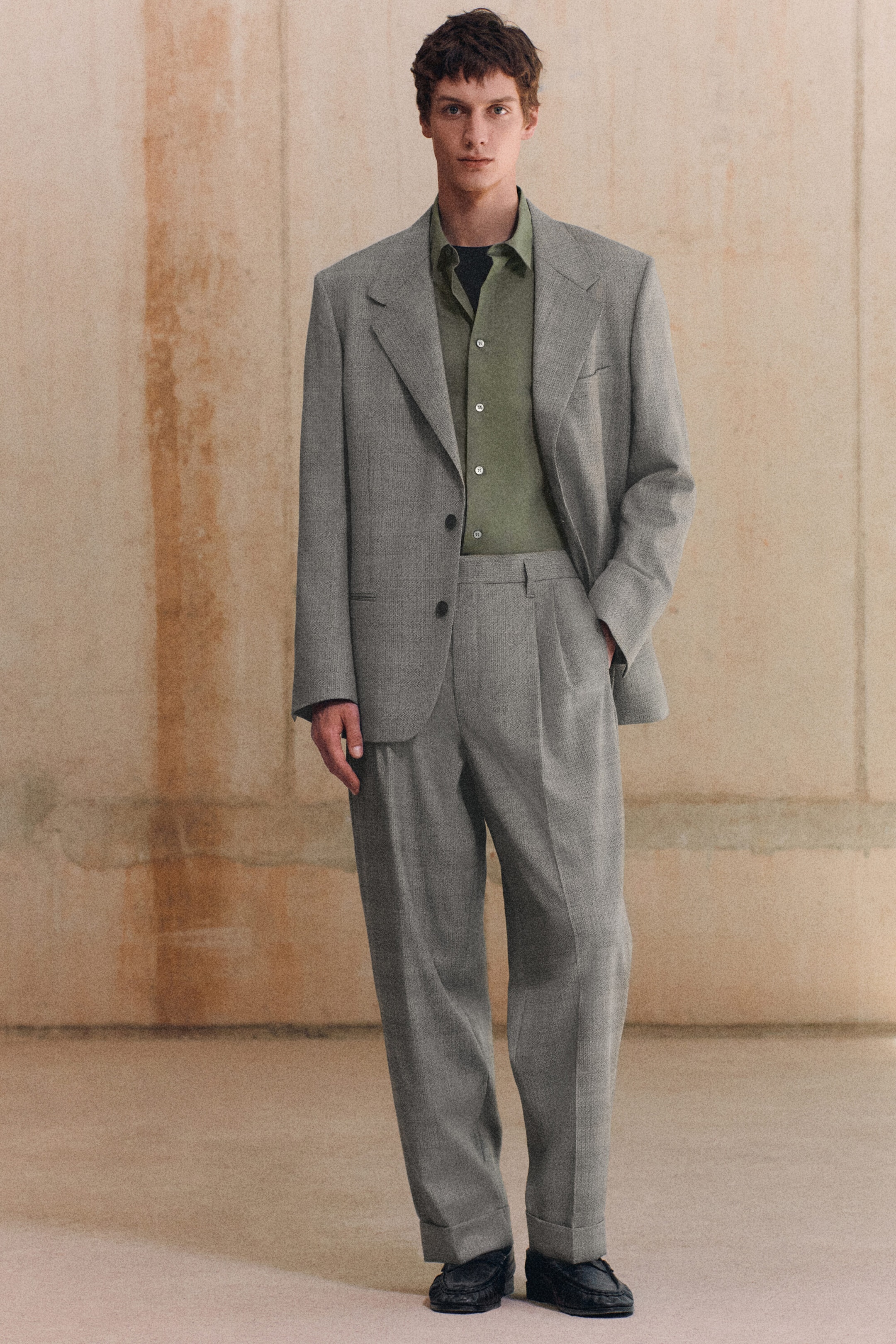 A model is facing the front, wearing a grey checked textured wool blazer and matching relaxed-fit tapered trousers. The single-breasted blazer features wide notch lapels and structured shoulders, while the trousers show front pleats and cuffed hems, layered over an olive green shirt. A model is facing the front, wearing a grey checked textured wool blazer and matching relaxed-fit tapered trousers. The single-breasted blazer features wide notch lapels and structured shoulders, while the trousers show front pleats and cuffed hems, layered over an olive green shirt.