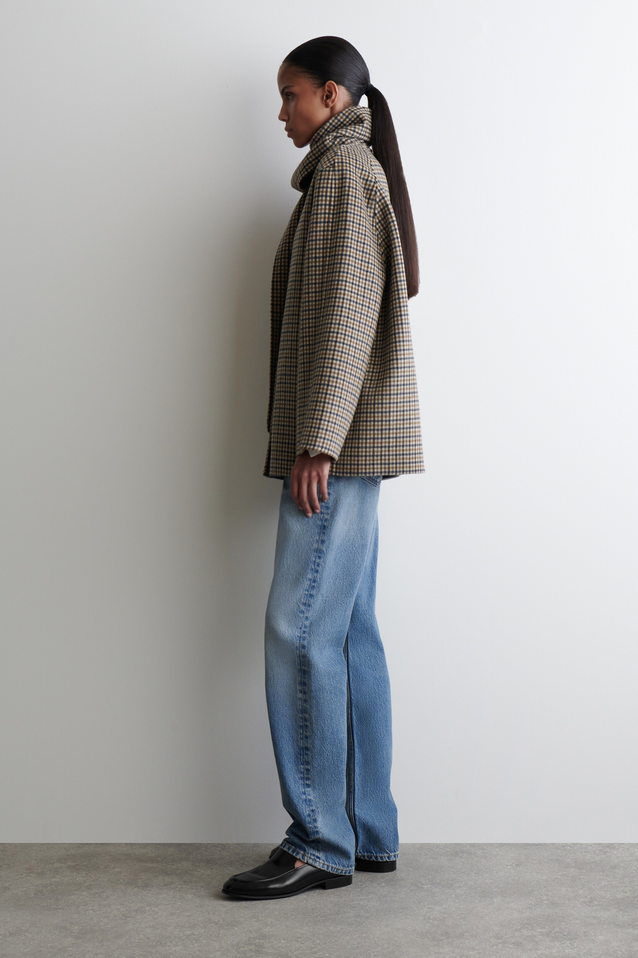 Größeres Bild anzeigen: Eine braune, schwarze und beige Hahnentrittjacke wird im Profil getragen, mit einem hohen, stehenden Kragen und einer lockeren, kastigen Passform. Sie wird mit hellblauen, weit geschnittenen Jeans kombiniert, die leicht über schwarzen Lederloafern aufliegen.