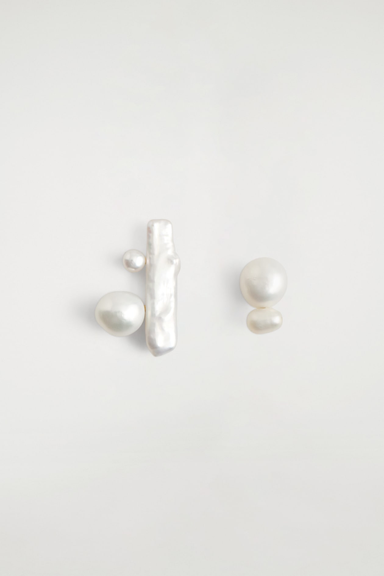 Sculptural freshwater pearl earrings are shown facing front, highlighting an asymmetrical design of iridescent white pearls. The left earring features a long, vertical baroque bar pearl flanked by a small and a medium round pearl, while the right earring displays a vertical stack of two round pearls of different sizes.