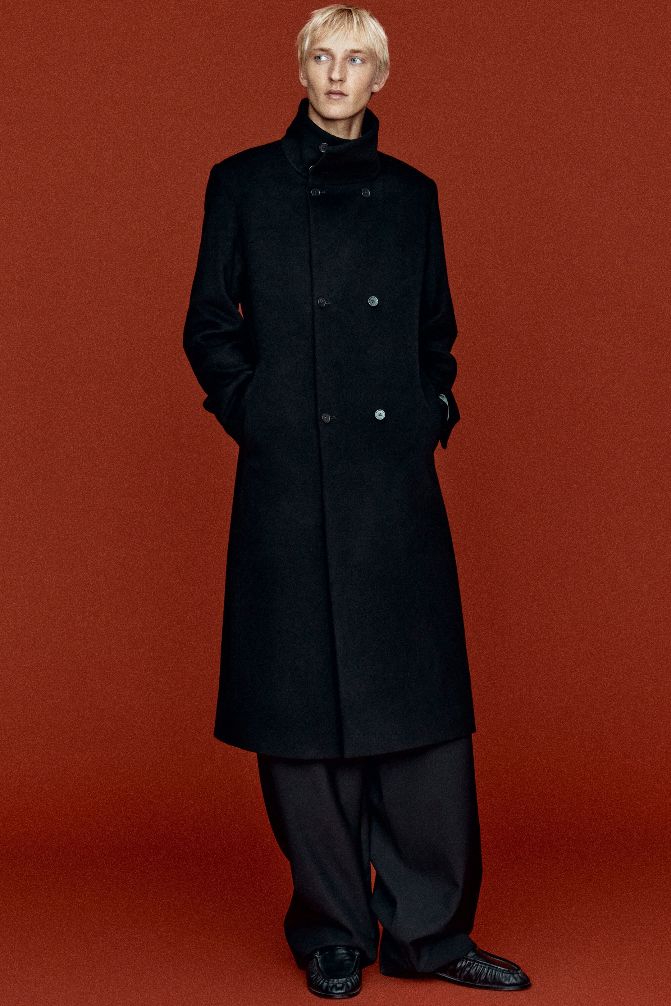 Un mannequin porte un long manteau en laine noir croisé, en vue de face, mettant en valeur sa silhouette structurée. Le manteau présente un col montant, des épaules marquées et une fermeture croisée à double boutonnage, et tombe sous le genou sur un pantalon noir à jambes larges. Un mannequin porte un long manteau en laine noir croisé, en vue de face, mettant en valeur sa silhouette structurée. Le manteau présente un col montant, des épaules marquées et une fermeture croisée à double boutonnage, et tombe sous le genou sur un pantalon noir à jambes larges.