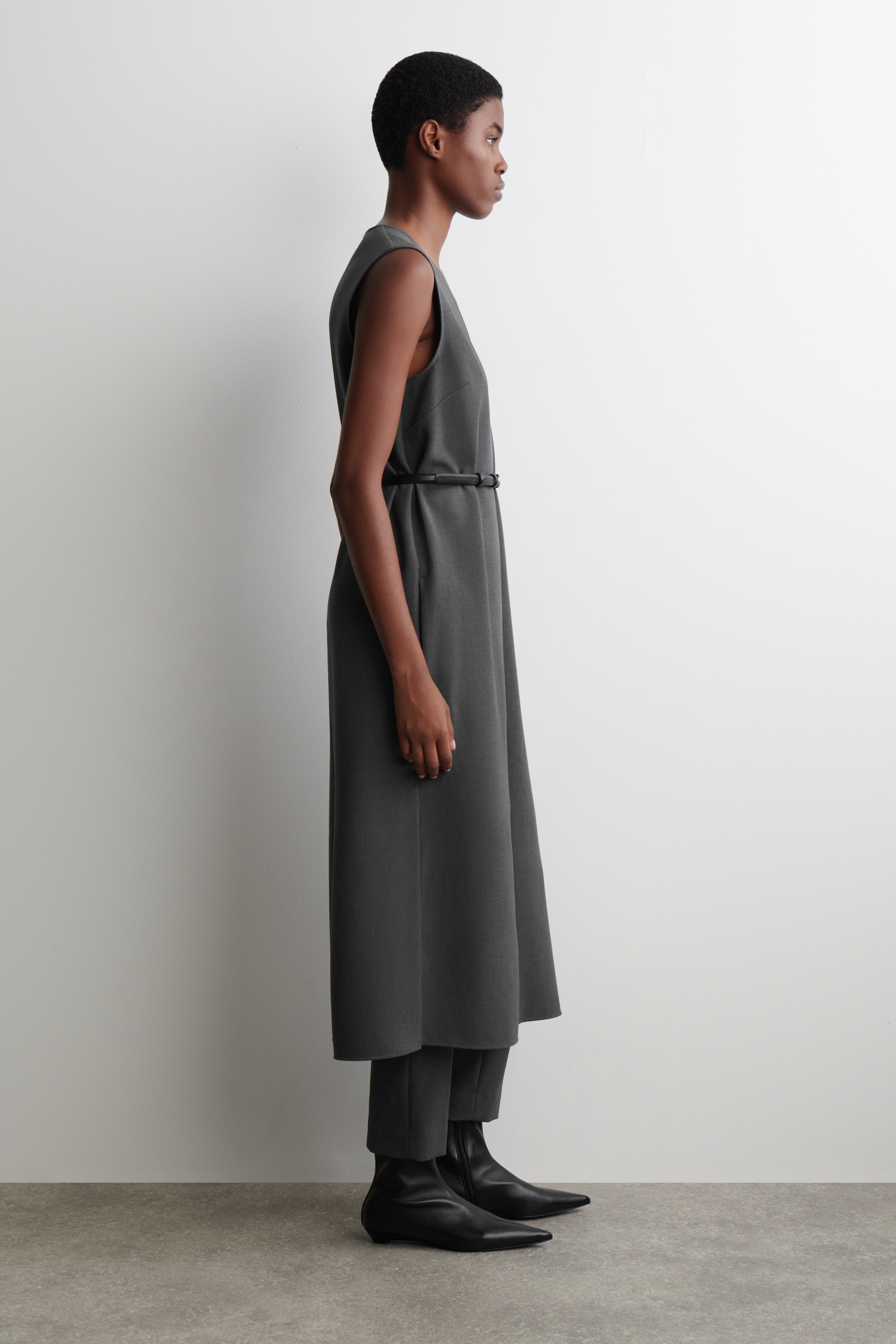 Agrandir l'image: Une robe midi sans manches gris foncé est présentée de profil, superposée à un pantalon court assorti. La robe présente un décolleté en V et est cintrée à la taille par une ceinture étroite foncée avec un accent métallique, complétée par des bottines noires à bout pointu.