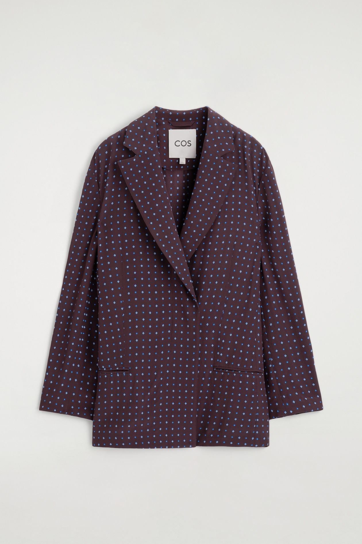 A front-facing double-breasted blazer with a diamond print showcases a dark brown fabric patterned with small blue and white geometric diamond motifs. The blazer features notch lapels, an overlapping front closure, and two horizontal welt pockets at the hips.