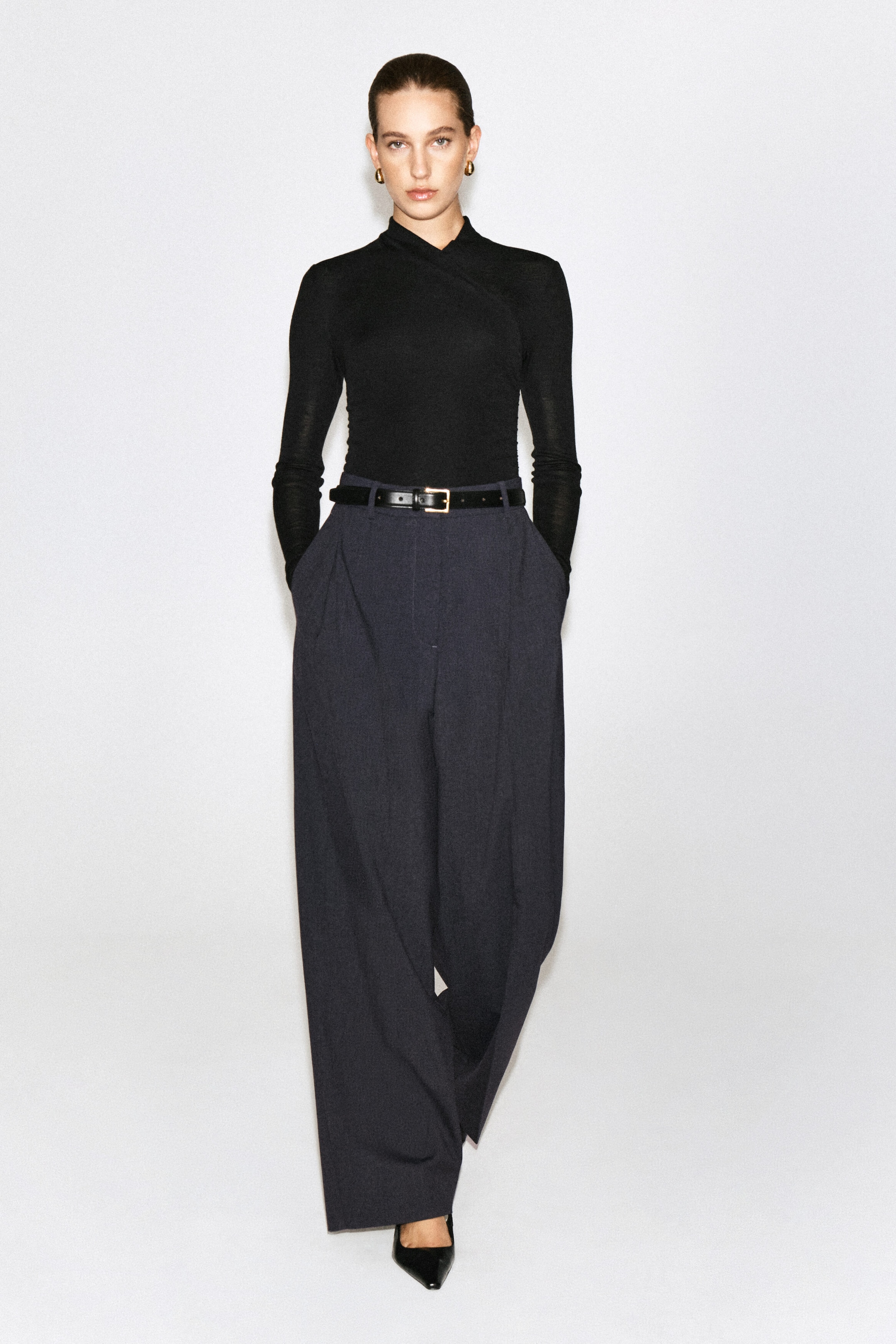 A black, slim-fit, long-sleeved top with a wrap-front shawl collar is being worn by the model. The top is tucked into wide-leg dark trousers styled with a black belt. A black, slim-fit, long-sleeved top with a wrap-front shawl collar is being worn by the model. The top is tucked into wide-leg dark trousers styled with a black belt.