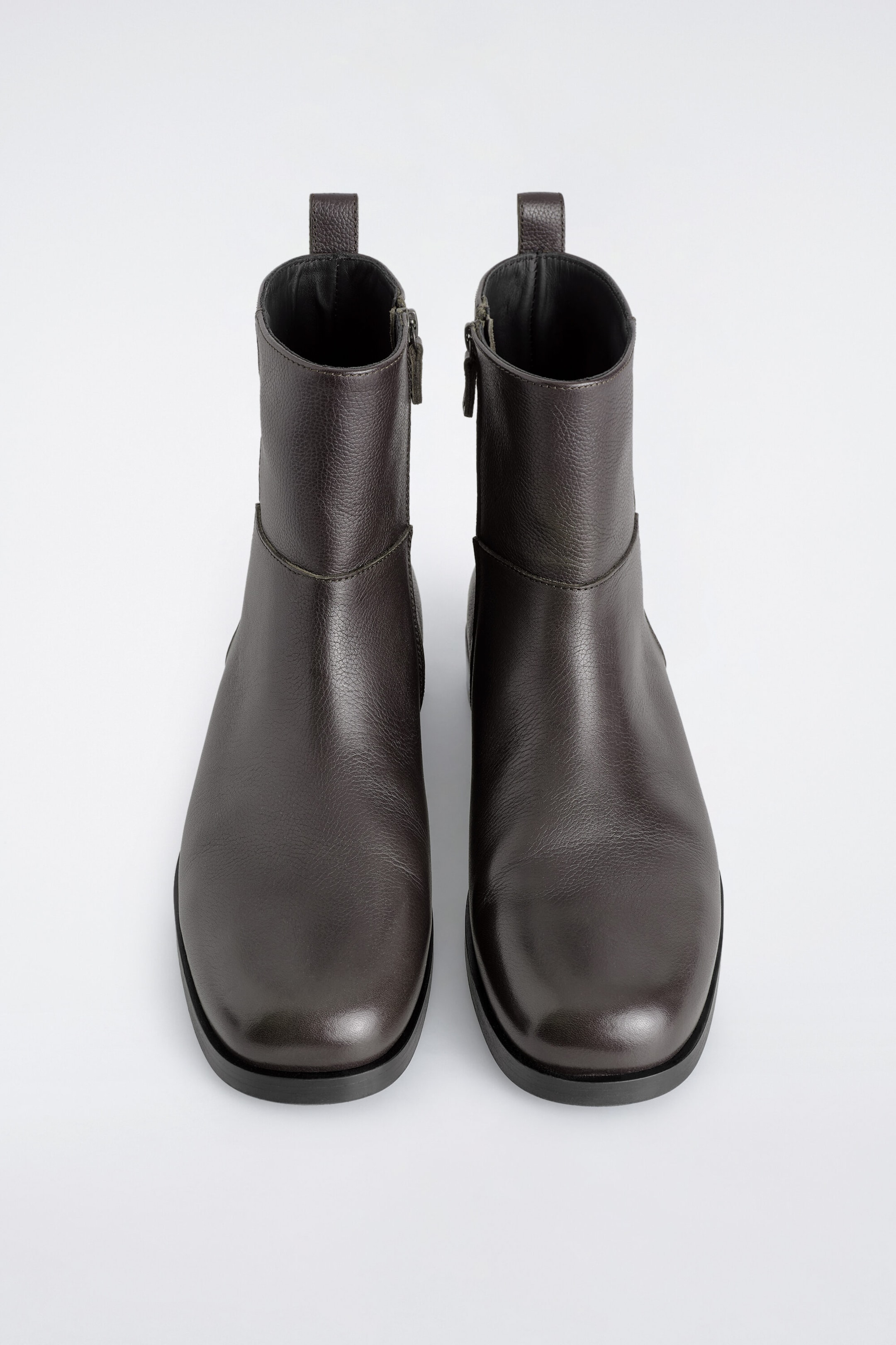 These square-toe leather ankle boots in dark brown stand upright, showcasing their textured grain leather and structured design from a top angle. They feature side zip fastenings, pull tabs at the back collar, and thick flat outsoles.