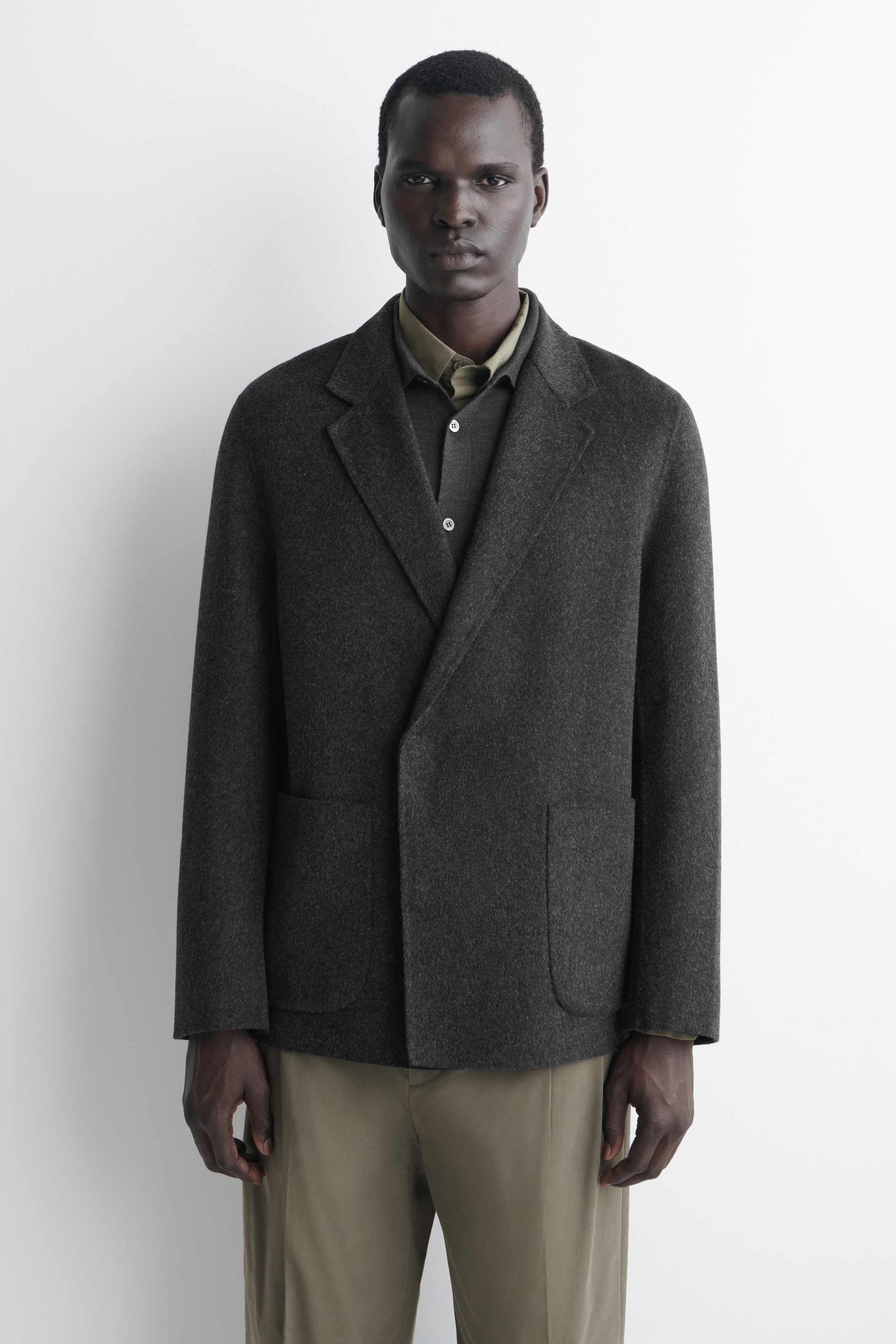 Ver imagen más grande: Se muestra de frente un blazer de mezcla de lana en gris oscuro, con un corte relajado y sin estructura, cierre de doble botonadura y solapas de muesca. El blazer presenta una tapeta frontal limpia y dos bolsillos de parche, combinado con una camisa de cuello en gris oscuro y pantalones verde oliva.
