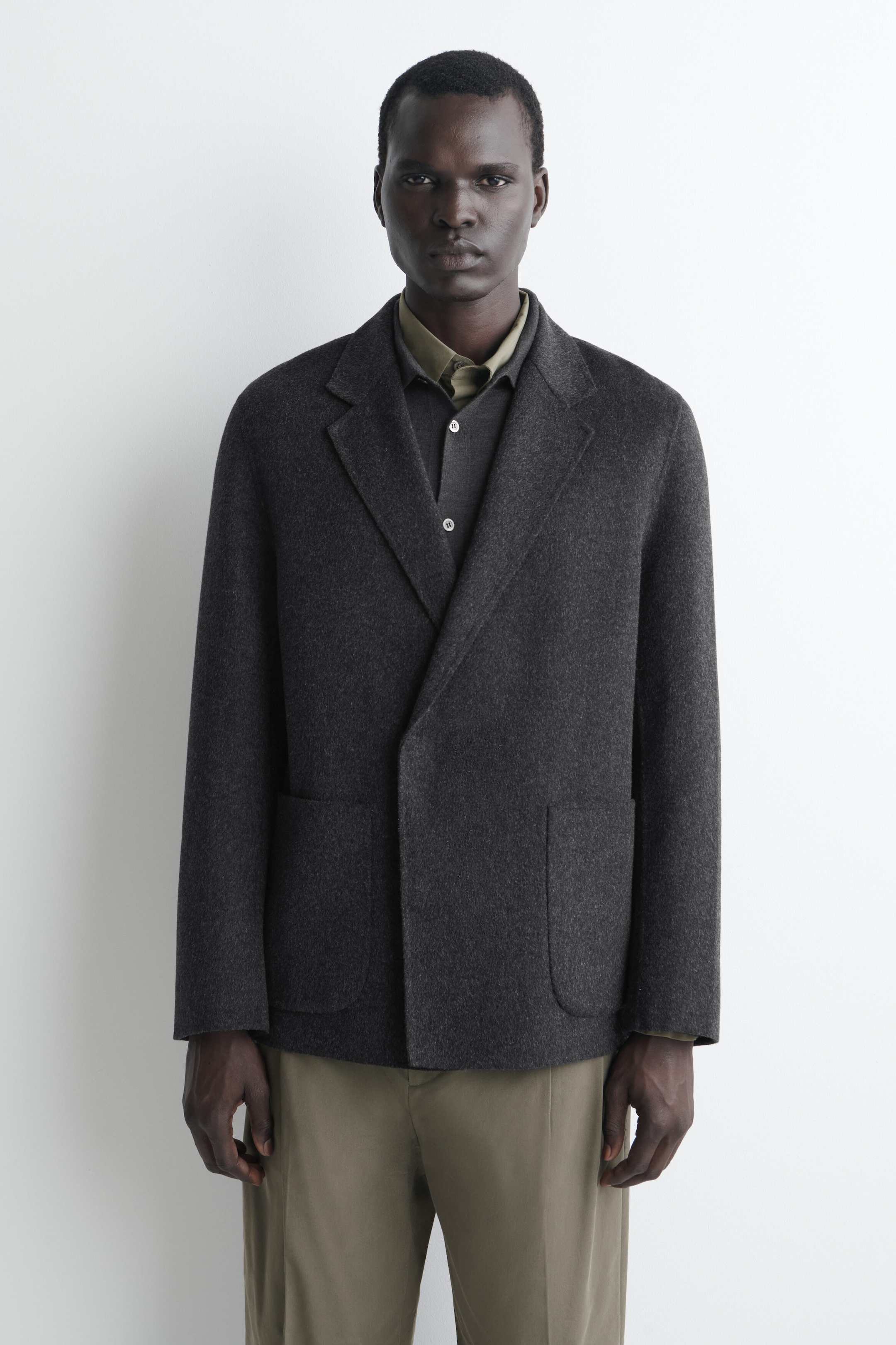 A dark grey wool-blend blazer is shown from the front, featuring a relaxed, unstructured fit with a double-breasted fastening and notch lapels. The blazer is designed with a clean front placket and two patch pockets, layered over a dark grey collared shirt and olive-green trousers. A dark grey wool-blend blazer is shown from the front, featuring a relaxed, unstructured fit with a double-breasted fastening and notch lapels. The blazer is designed with a clean front placket and two patch pockets, layered over a dark grey collared shirt and olive-green trousers.