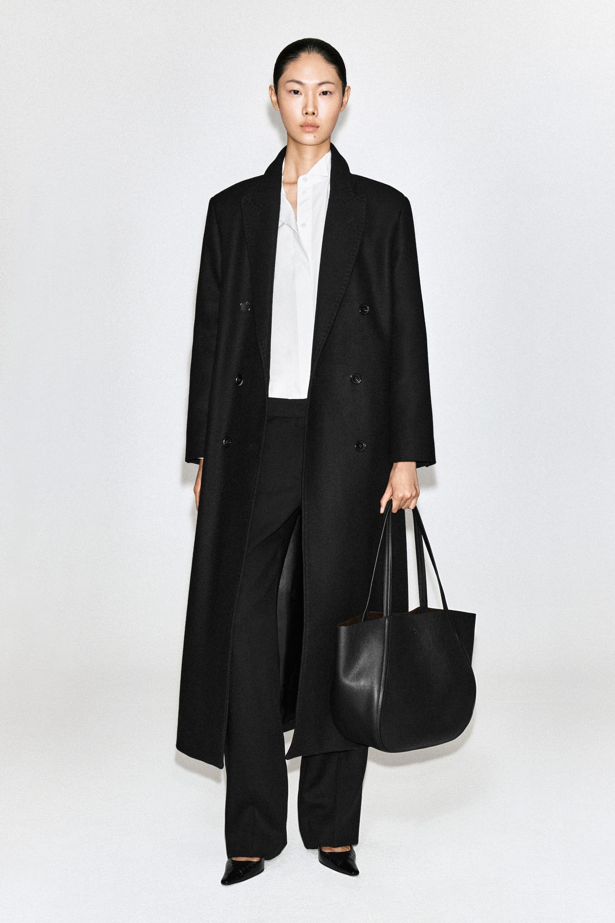 A black leather shopper bag with a curved silhouette and dual top handles is being carried by the model at her side. The model is wearing a long double-breasted black coat, a white collared shirt, and black trousers. A black leather shopper bag with a curved silhouette and dual top handles is being carried by the model at her side. The model is wearing a long double-breasted black coat, a white collared shirt, and black trousers.