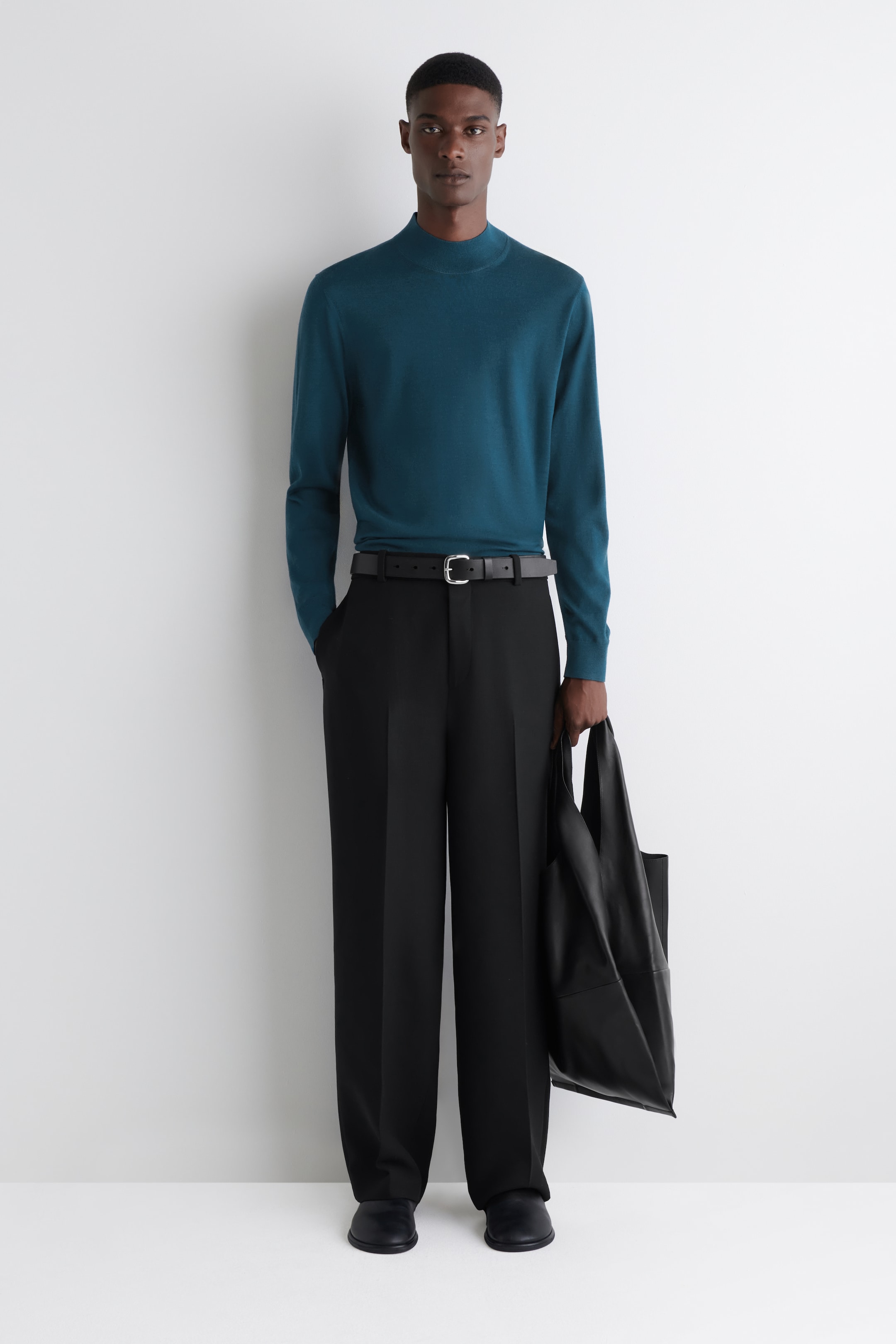 View larger image: A long-sleeved, teal mock neck sweater is being worn tucked into black wide-leg trousers, which are cinched with a black leather belt featuring a silver-toned buckle. Black flat shoes are visible, and a black leather tote bag is being held in one hand.