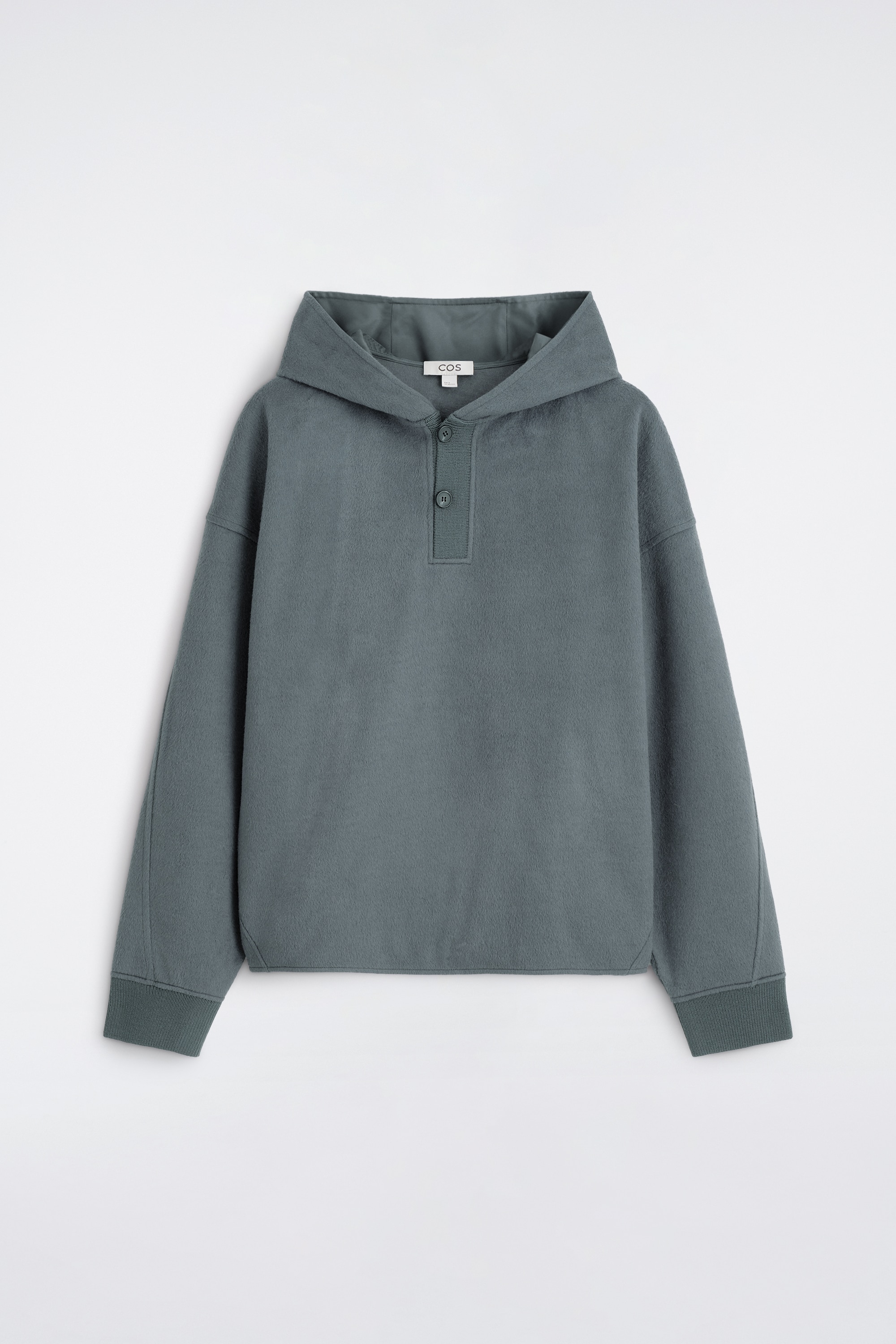 Un pull Ã  capuche vert grisÃ¢tre avec encolure henley boutonnÃ©e et poignets cÃ´telÃ©s est portÃ© par le mannequin. Le haut Ã  coupe dÃ©contractÃ©e est superposÃ© Ã  un t-shirt blanc et associÃ© Ã  un jean bleu.