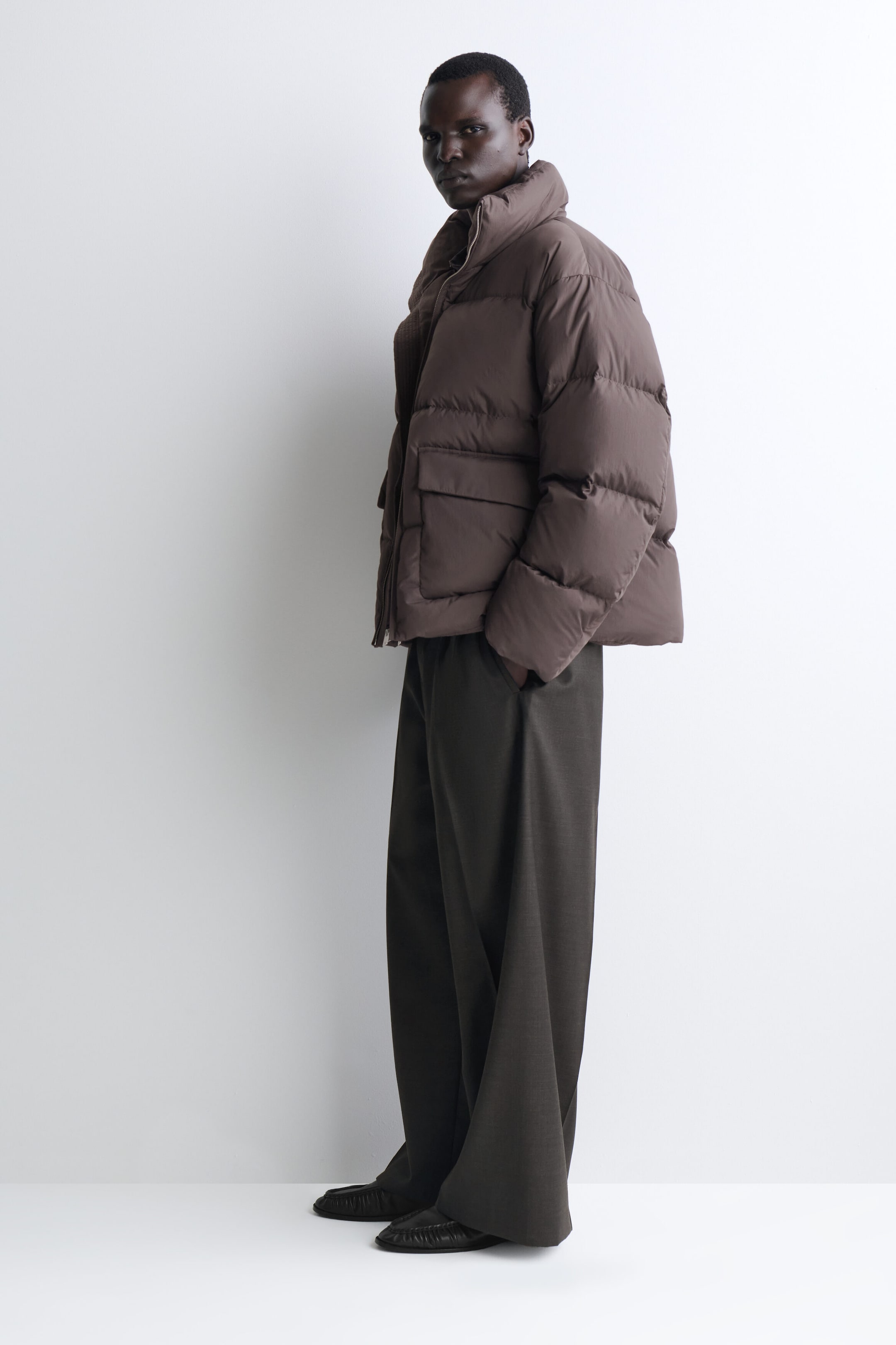 A brown funnel-neck nylon down puffer jacket is shown facing left, highlighting its voluminous quilted design, high collar, and large front flap pockets. The jacket is styled with dark, wide-leg trousers to emphasize its relaxed, oversized fit. A brown funnel-neck nylon down puffer jacket is shown facing left, highlighting its voluminous quilted design, high collar, and large front flap pockets. The jacket is styled with dark, wide-leg trousers to emphasize its relaxed, oversized fit.